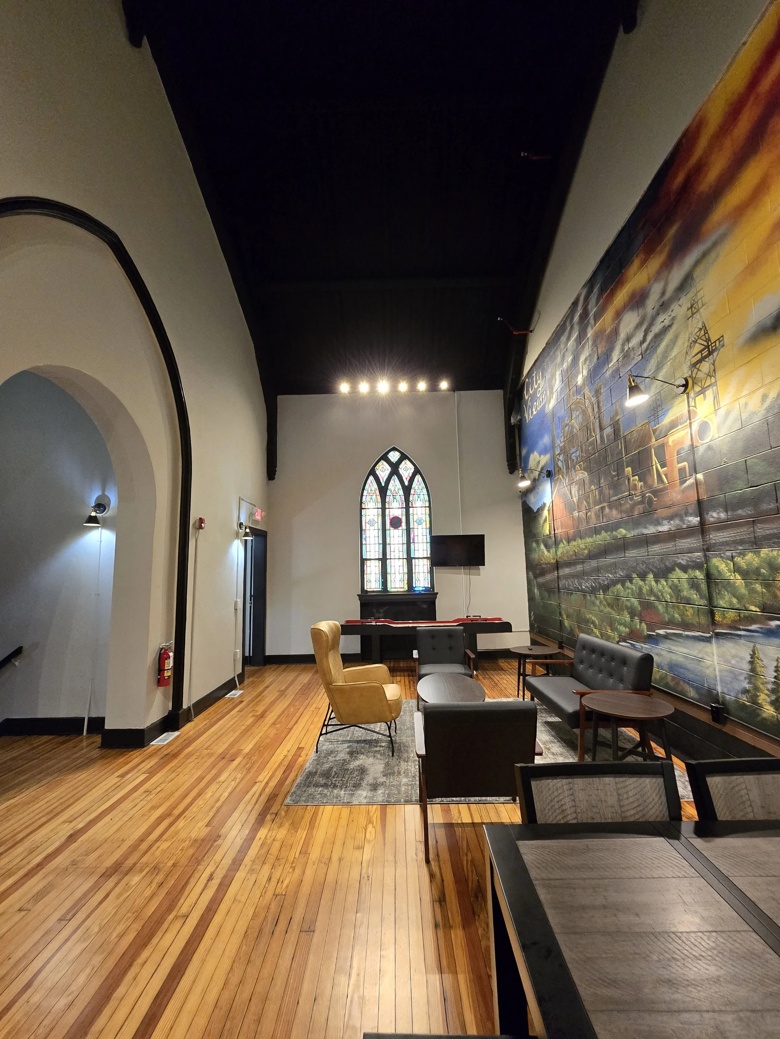 Interior of a spacious room with wooden floors, a large mural on the right wall, and a stained glass window at the back. Furniture includes chairs, a small table, and a TV mounted on the wall.