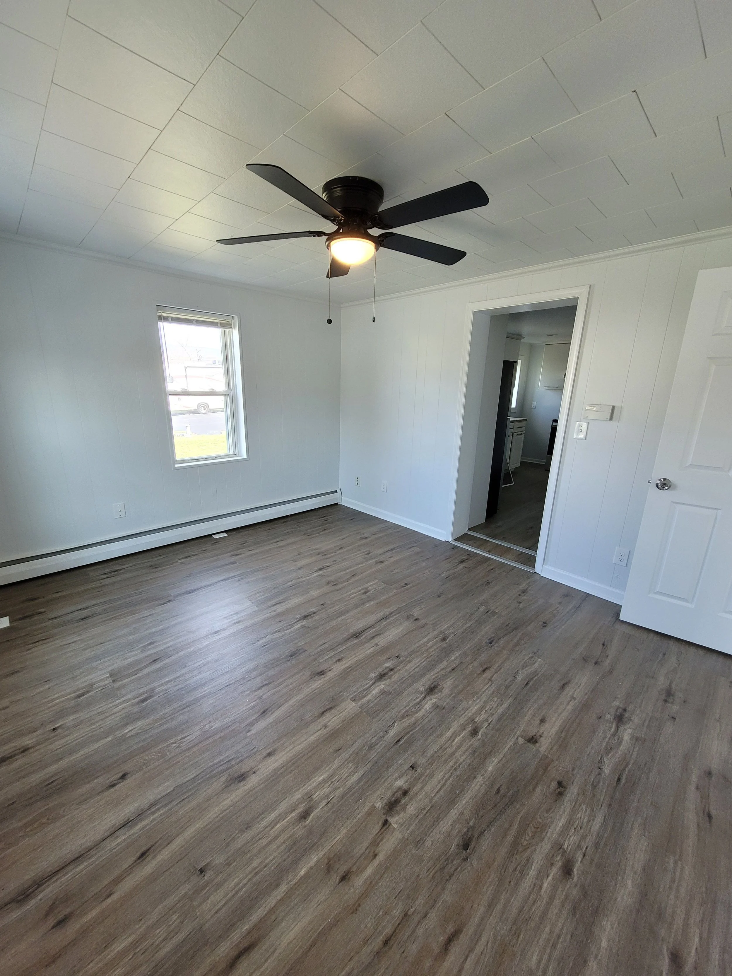 Empty room with wood flooring, white paneled walls, a window, a ceiling fan with light, and a doorway leading to another room.