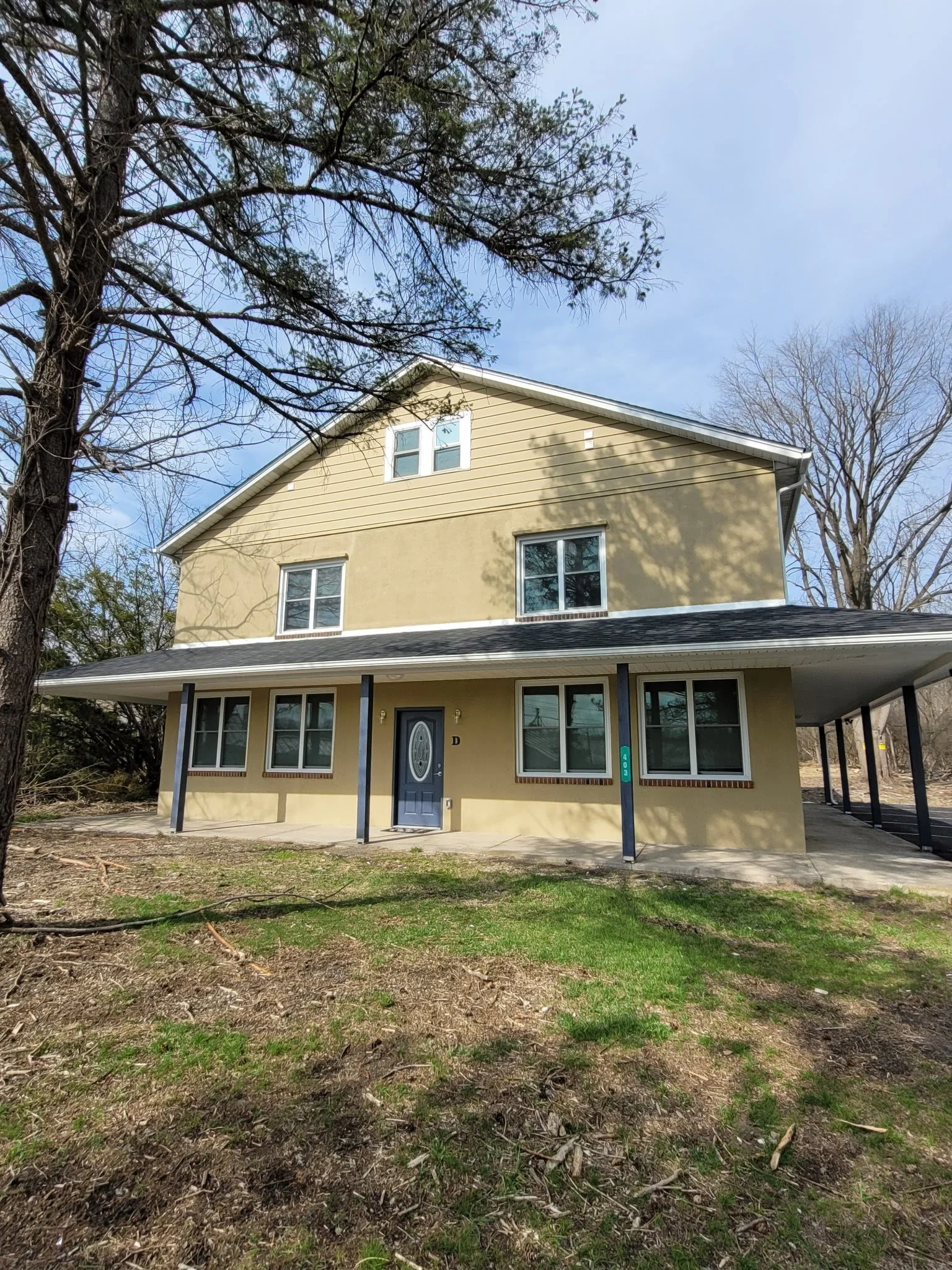 A two-story beige house with multiple windows, a dark blue front door, and a porch supported by black columns, situated on a grassy yard with trees and a partly cloudy sky.