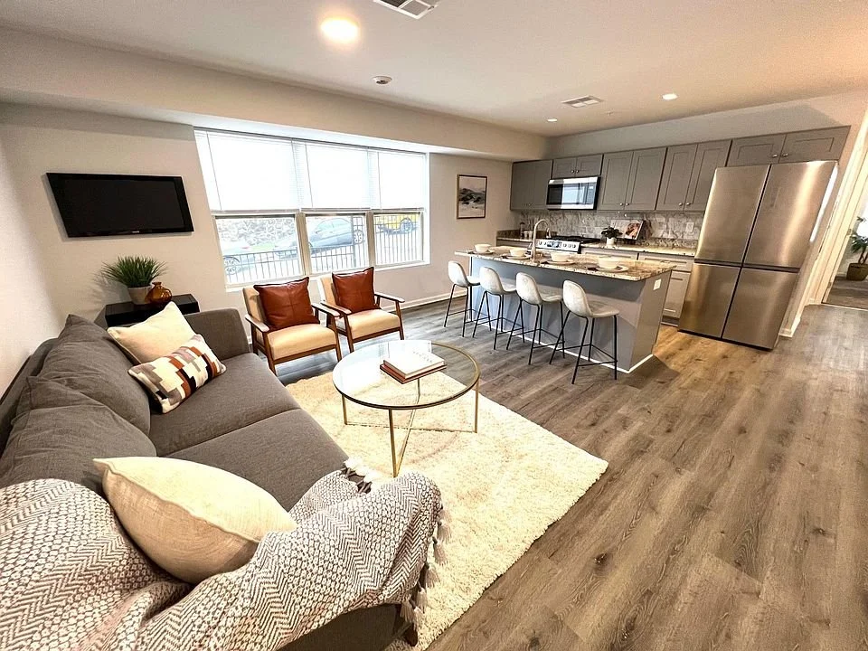 Open-concept living room and kitchen with gray sofa, two armchairs, coffee table, wall-mounted TV, kitchen island with four chairs, gray cabinets, stainless steel refrigerator, and window with blinds.