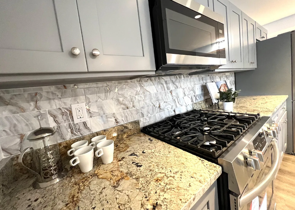 Kitchen countertop with coffee plunger, three small white cups, stove with four burners, microwave above, gray cabinets, and a decorative plant.