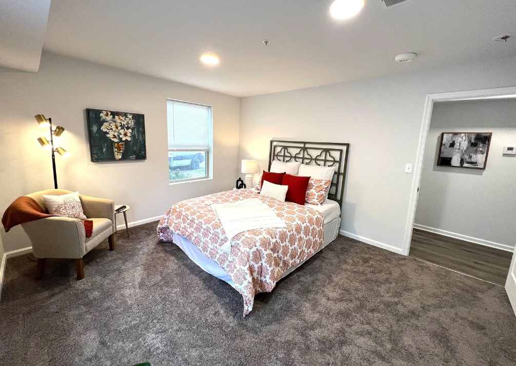 Bedroom with a bed, pillows, floral bedding, a nightstand, a lamp, an armchair with a blanket and pillow, a painting, a window, and a doorway leading to another room.