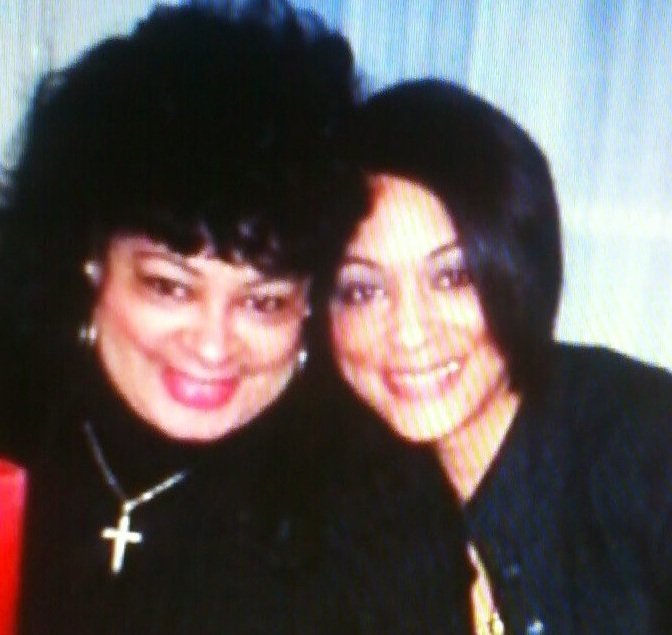 Two women smiling closely together, one with dark, voluminous hair and wearing a cross necklace, the other with straight dark hair, both dressed in dark clothing.