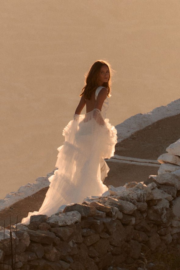 Woman in a flowing white wedding dress standing on a rocky stone terrace at sunset