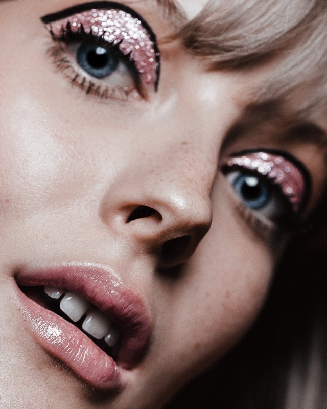 Close-up beauty photograph of a face with theatrical eye makeup featuring pink and silver glitter eyeshadow, black winged eyeliner, and glossy mauve lips.
