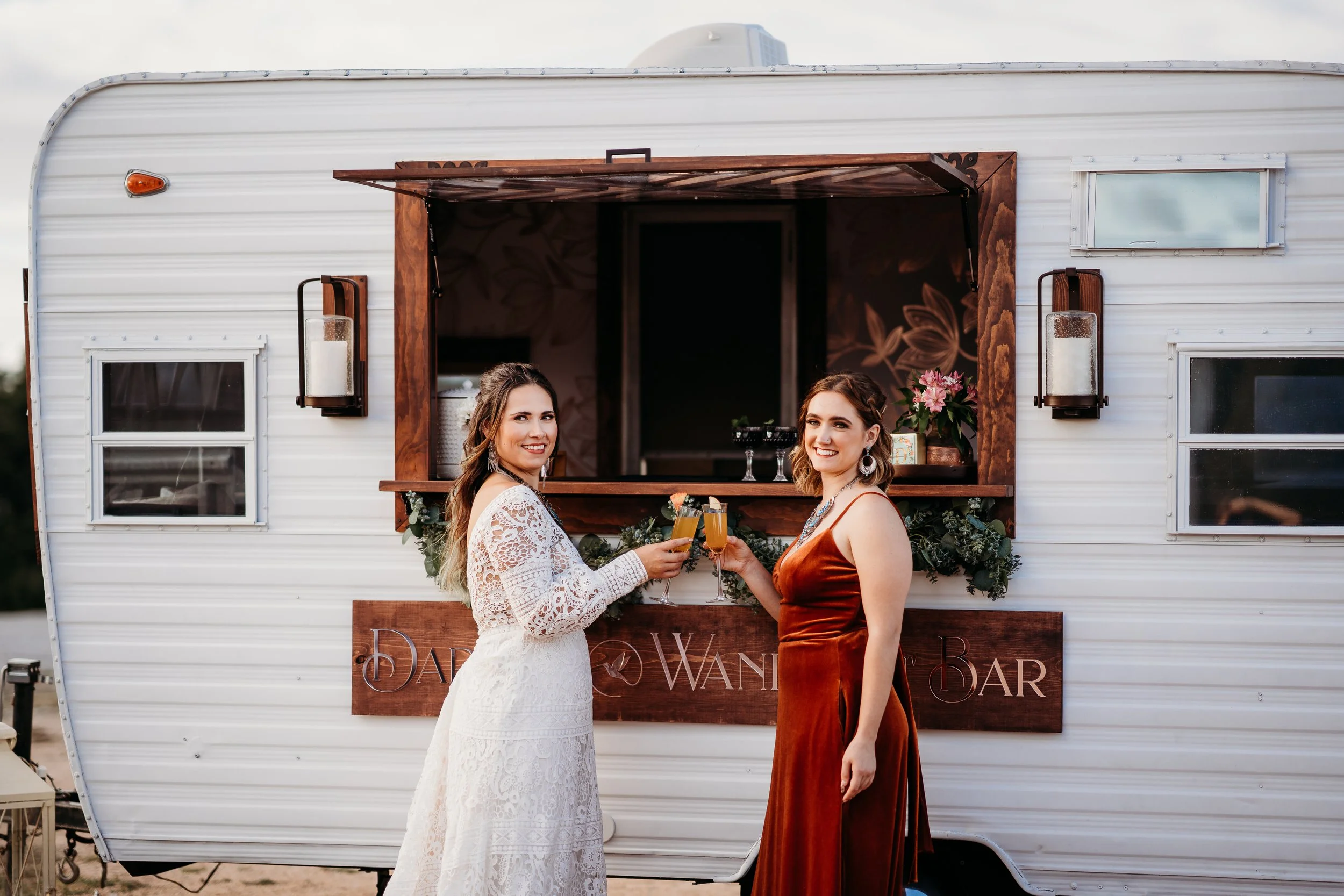 Bride and bridesmaid toasting with champagne at custom mobile bar during a Camp Hideaway wedding in Spicewood, Texas