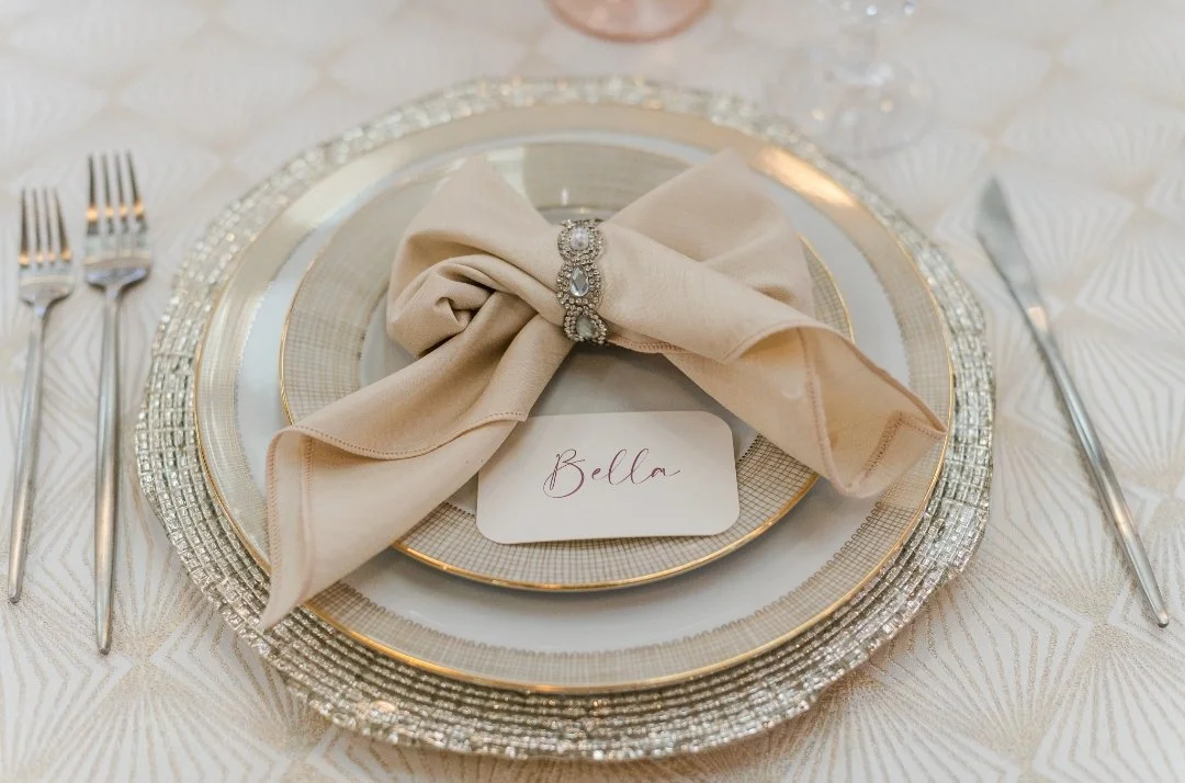 Elegant wedding place setting featuring layered charger plates, a soft champagne linen bow napkin, and a handwritten place card.