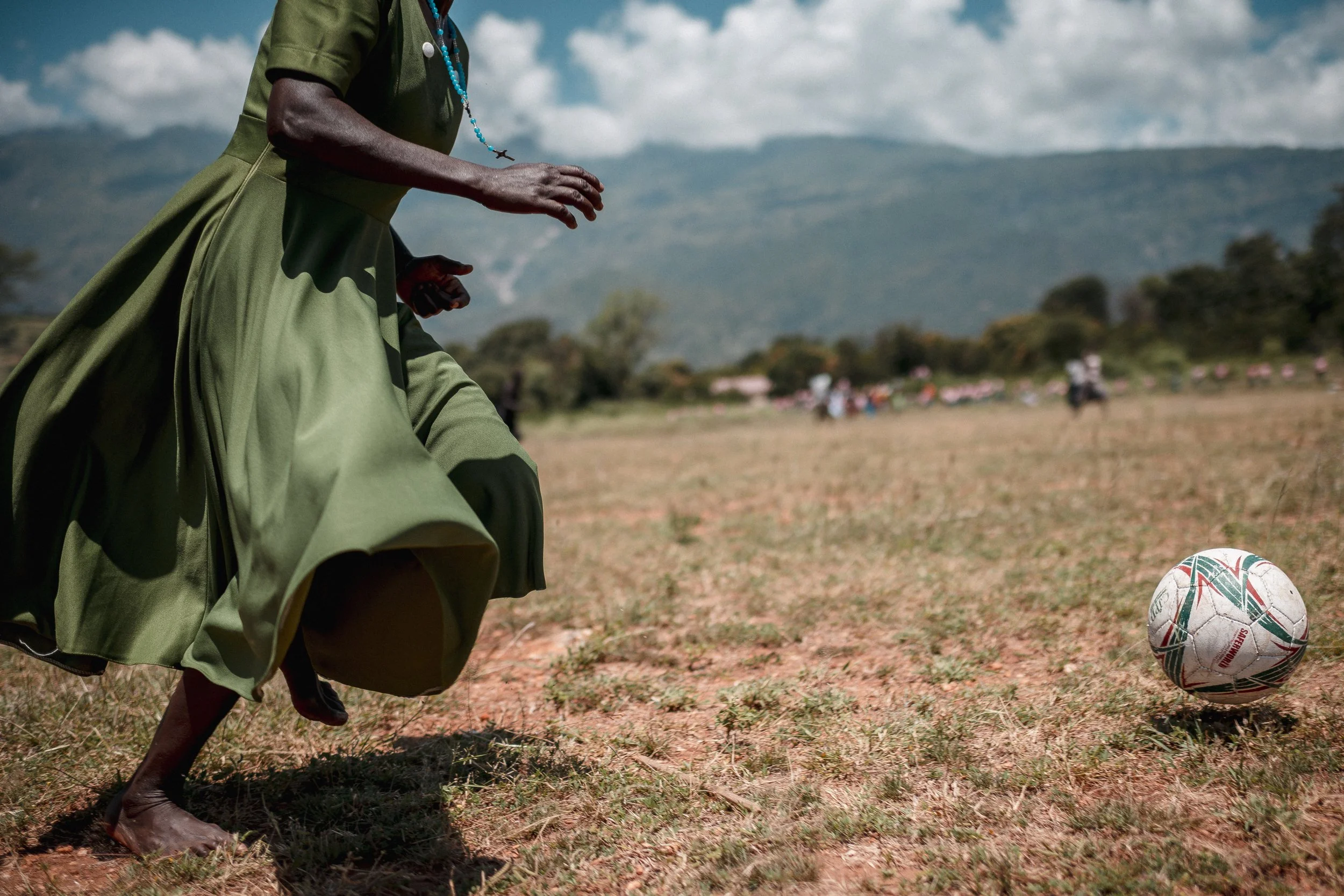A person wearing a green dress and barefoot is about to kick a soccer ball on a dry grassy field. In the background, there are mountains and a group of people.
