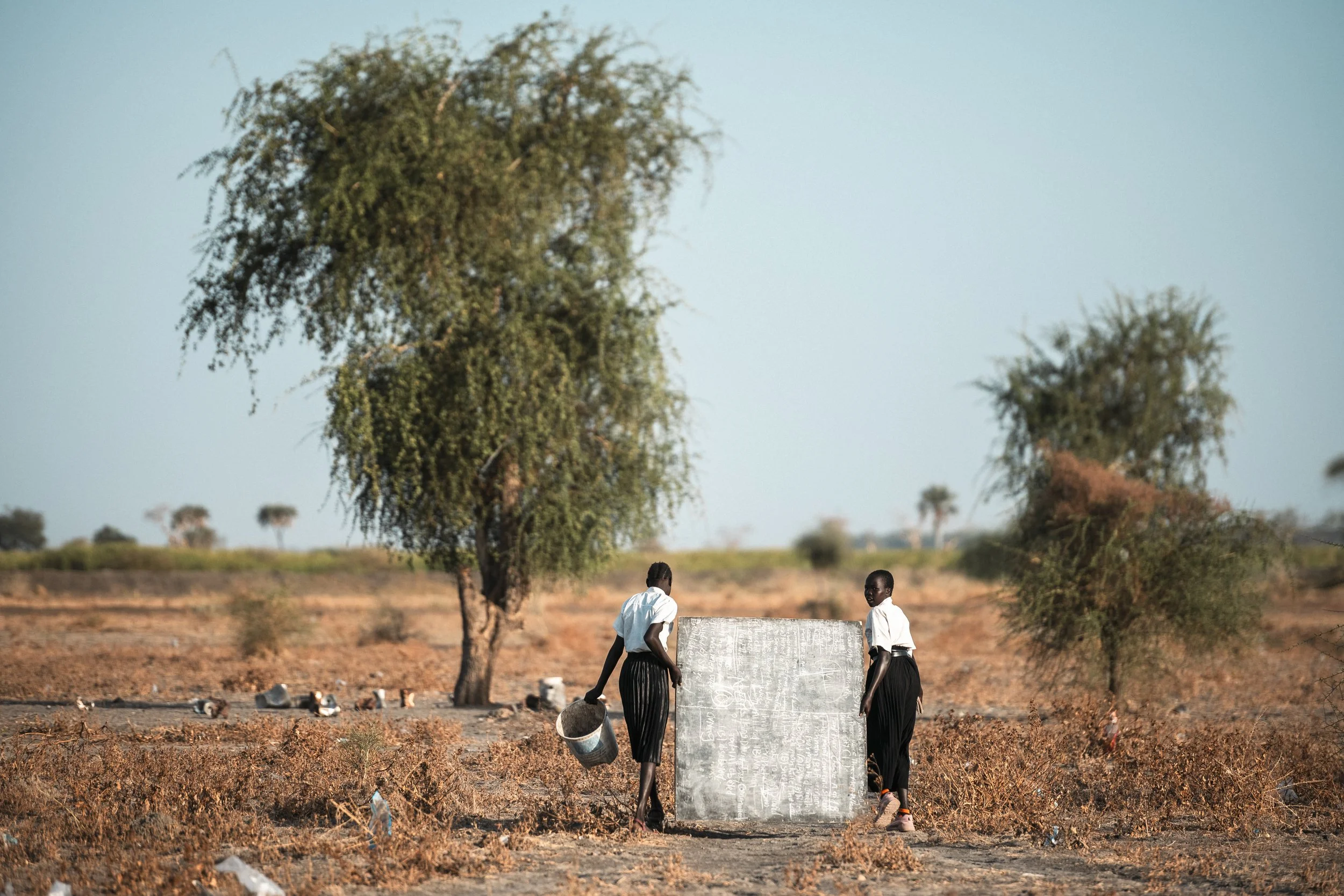 Two girls carry a chalkboard across the dry ground to set up their classroom beneath a tree in New Fangak.