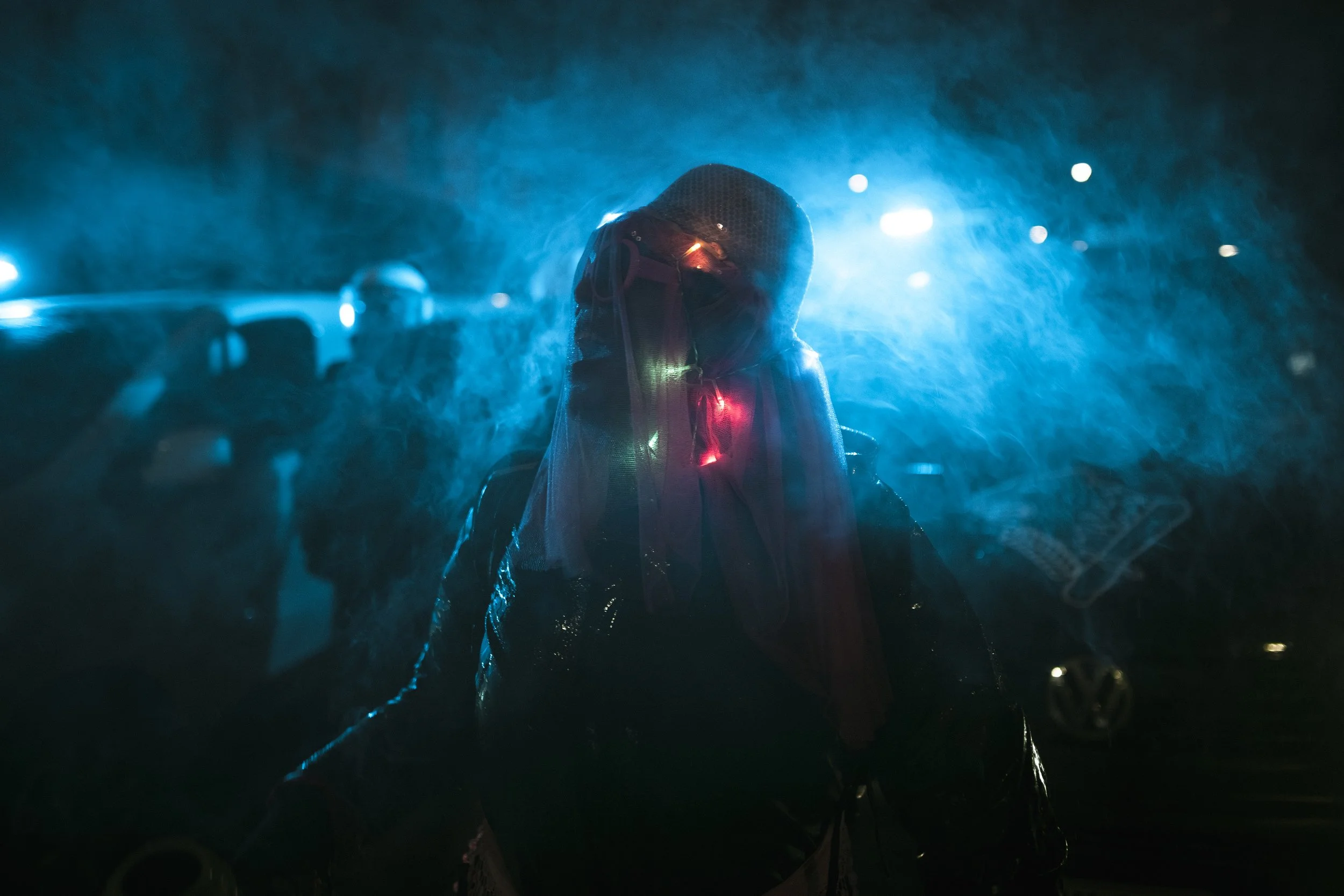 Person standing in the rain at night, illuminated by blue and red lights, with visible water droplets and vehicles in the background.