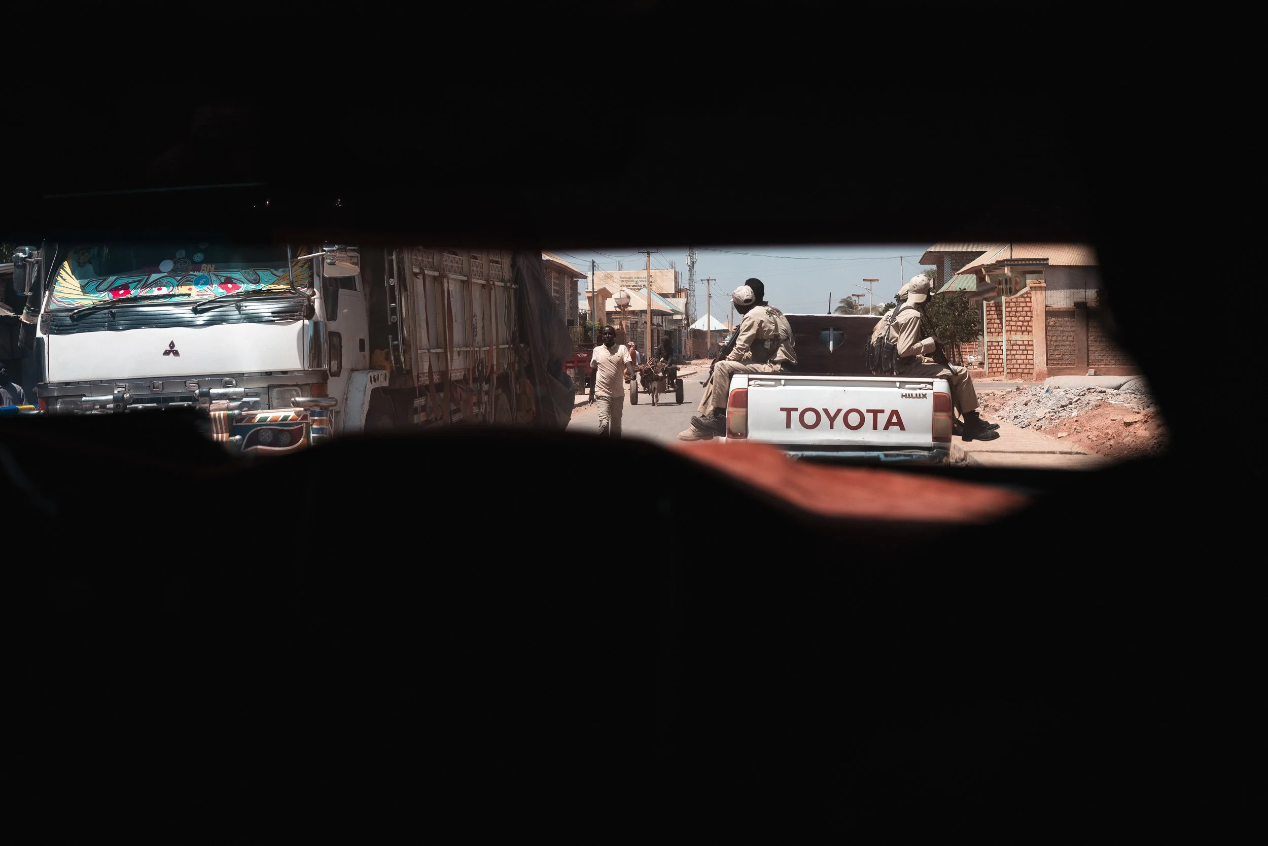 View through a vehicle window showing a street scene with a Toyota Hilux pickup truck carrying uniformed men sitting on the back, a few buildings, and some pedestrians in the background.