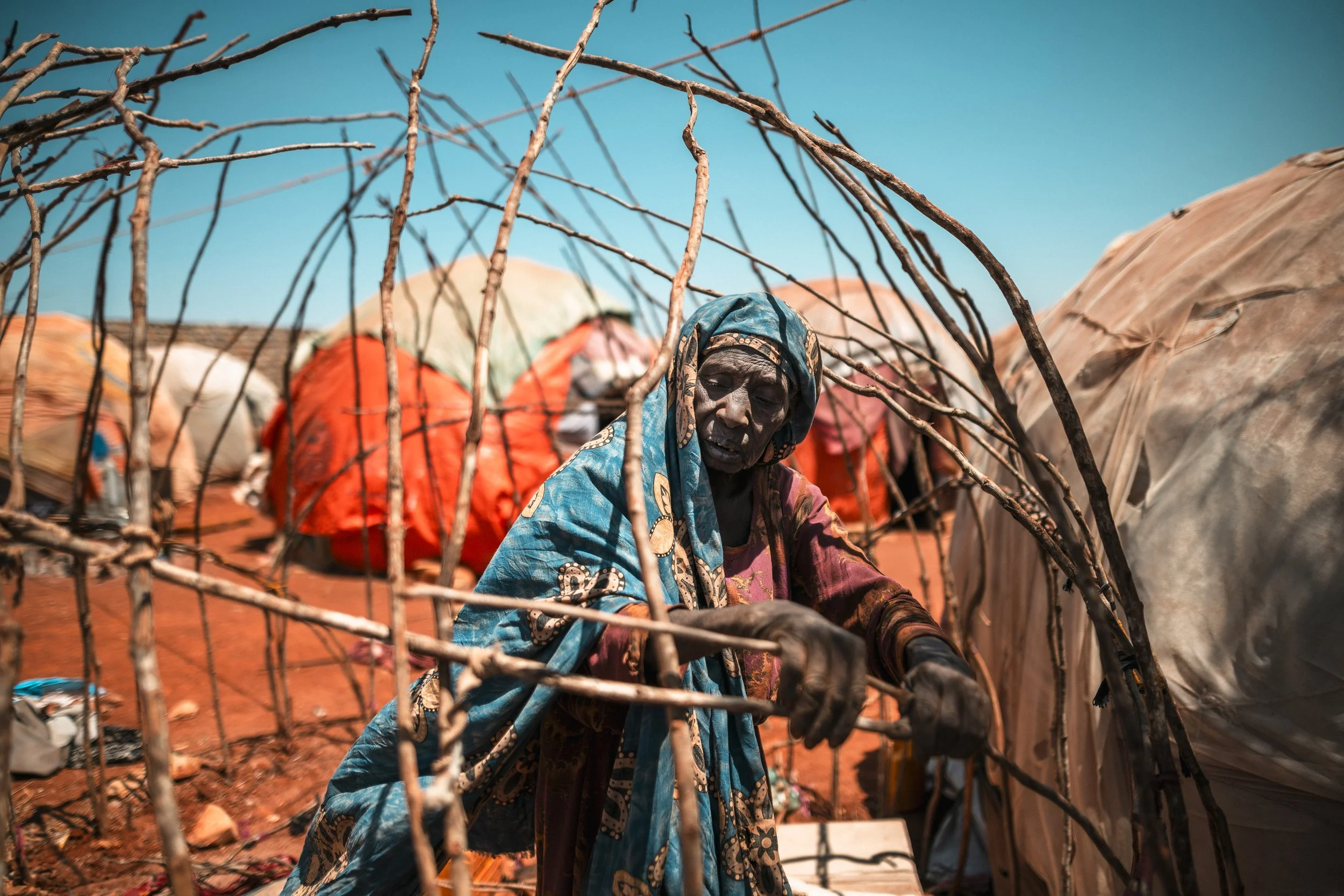 An elderly woman in colorful traditional clothing working outdoors, surrounded by wooden sticks and fabric-covered structures, with plenty of bright sunlight and a clear blue sky.