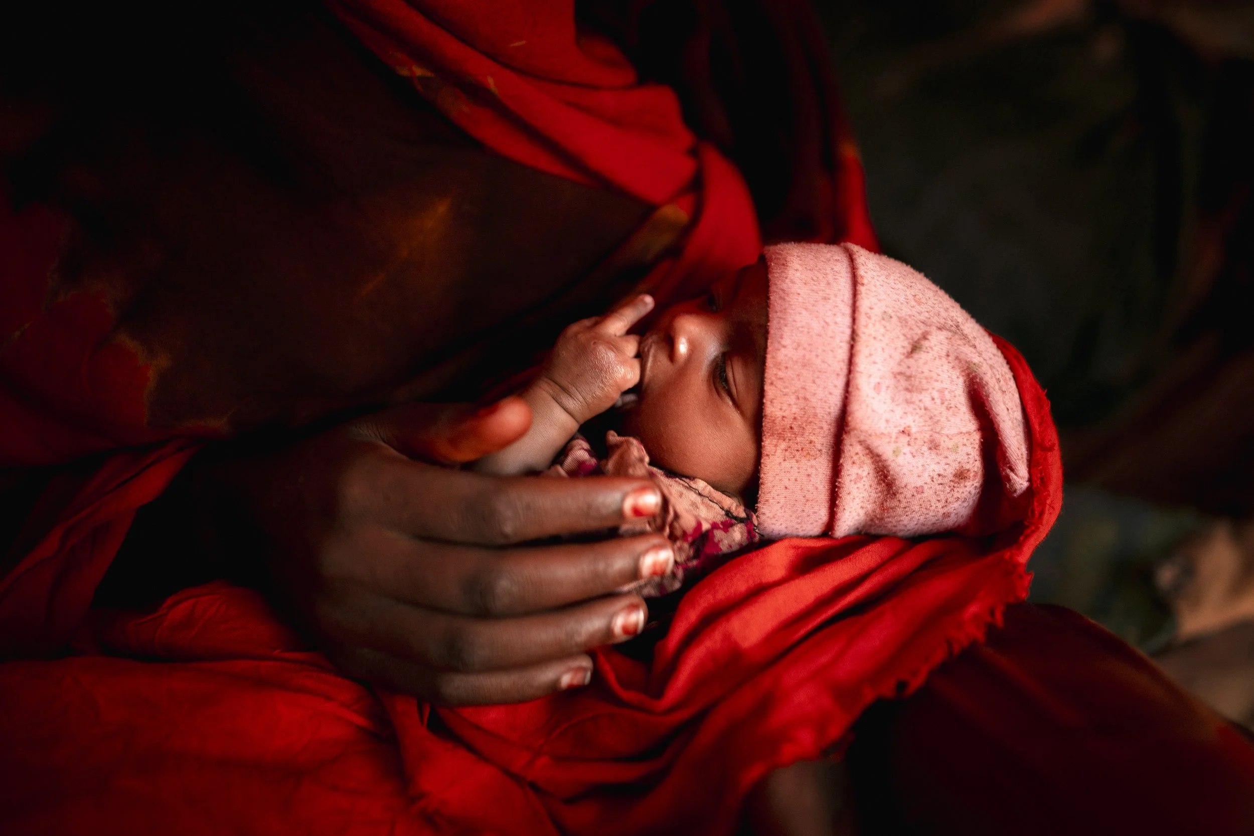 A close-up of a person holding a newborn baby wrapped in red cloth. The baby is wearing a pink hat and sucking on their finger, with the person's hand gently supporting the baby's head and body.