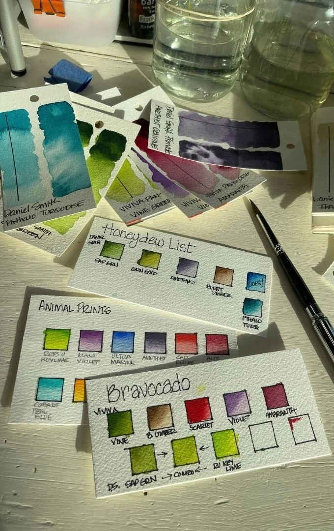 Hand-painted watercolor swatches in turquoise, green, violet, red, and blue fill labeled test cards for WhimZadoodles artwork, with a paintbrush and water jar nearby.