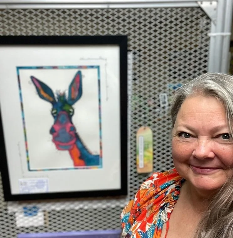 Jake Derr, the artist behind WhimZadoodles, standing in front of her colorful watercolor artwork, Clementine Cloverbottom, and its Honorable Mention award.