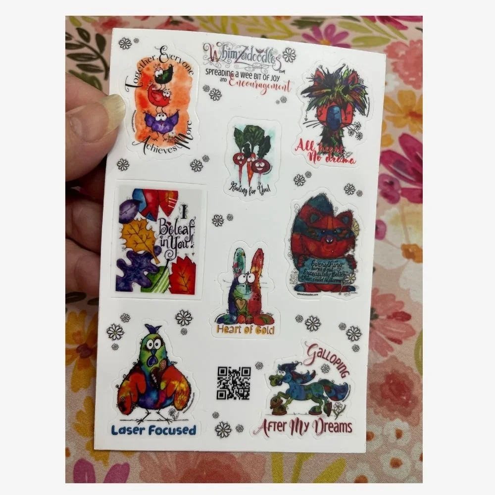 Hand holding a whimsical watercolor encouragement sticker sheet featuring inspirational quotes and colorful characters, showing the size and detail of the stickers.