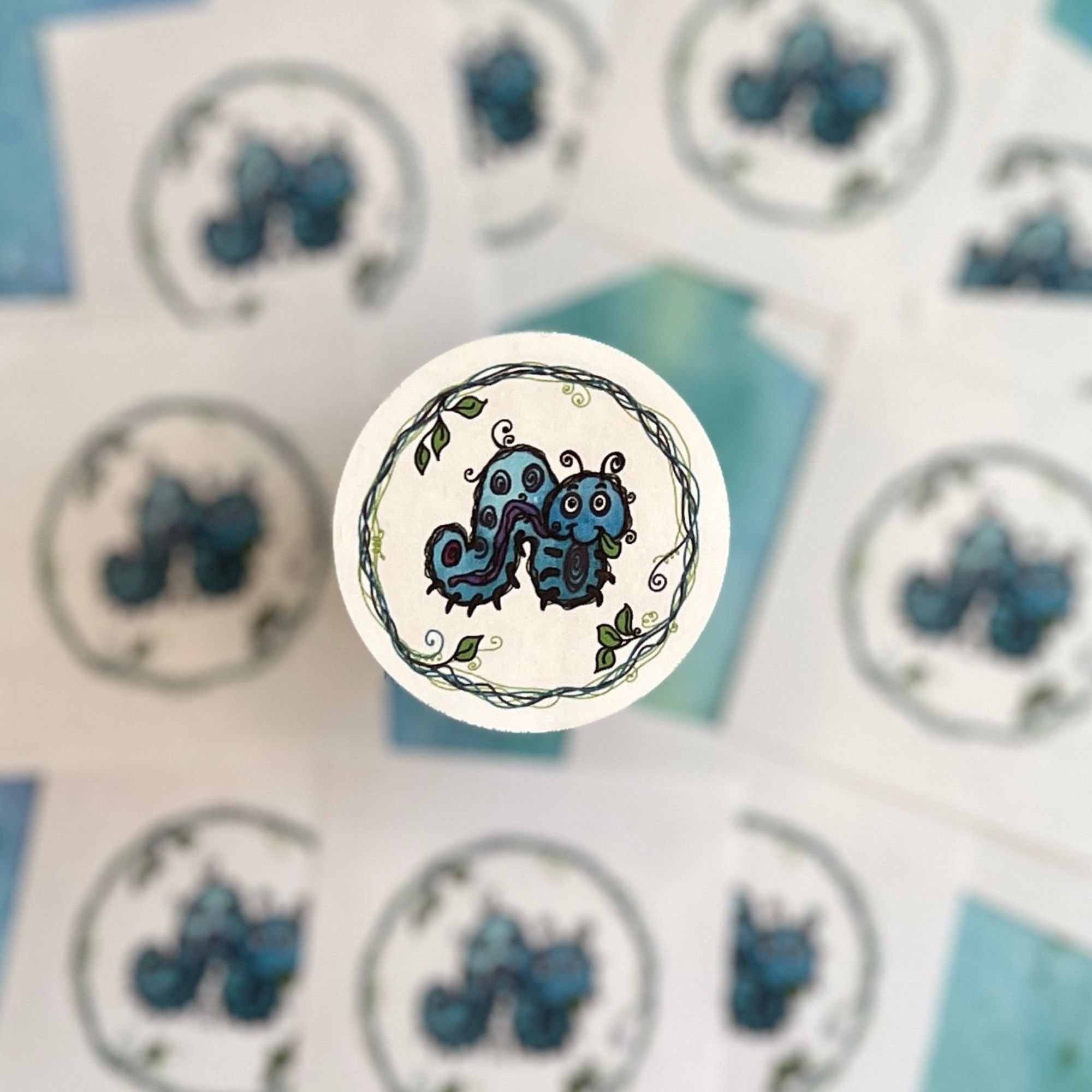 Close-up of Munchie caterpillar envelope seal showing playful swirl patterns, smiling face, and leaf in mouth with decorative vine border