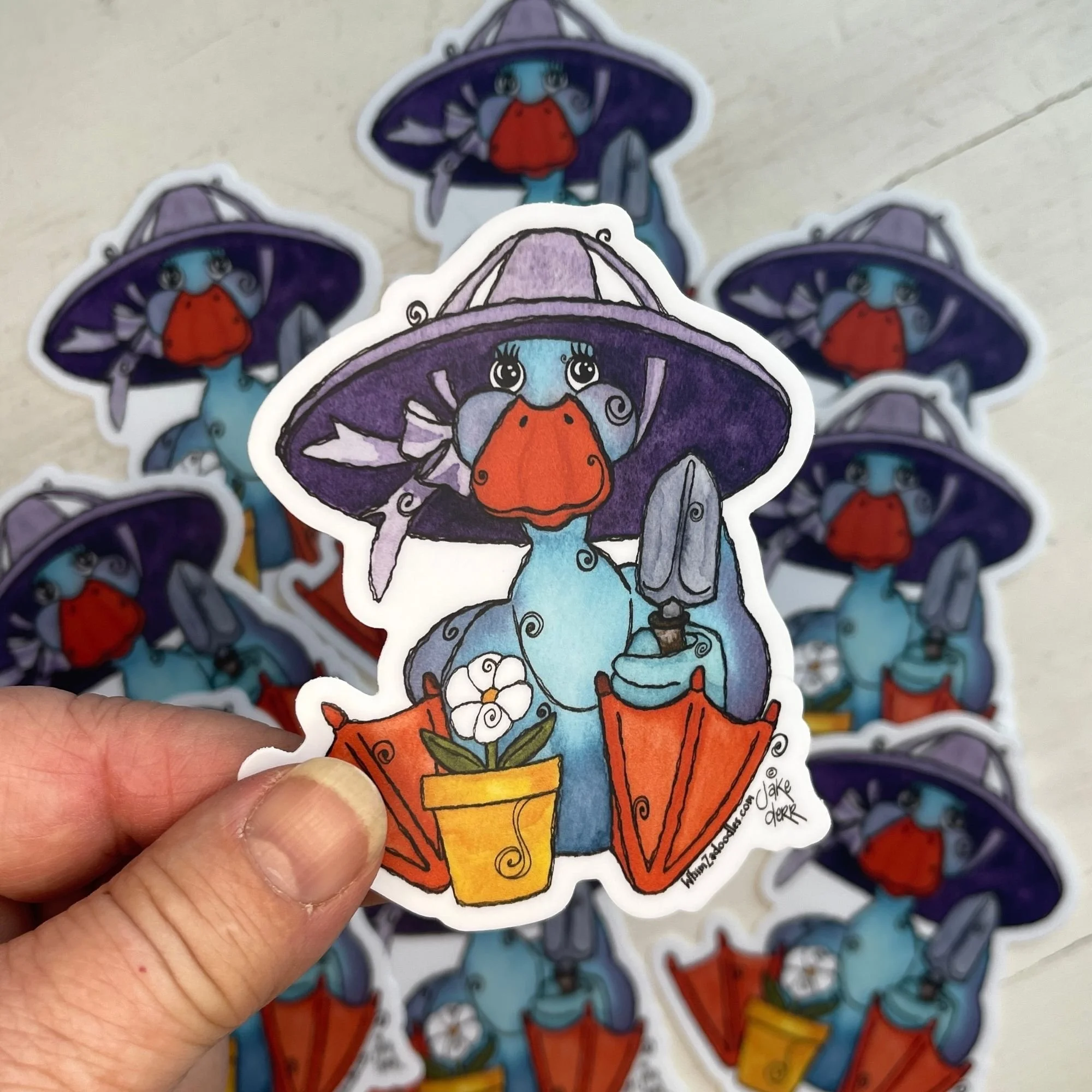 Hand holding a teal duck gardening sticker with purple sun hat and trowel, foreground in focus with softly blurred neutral background