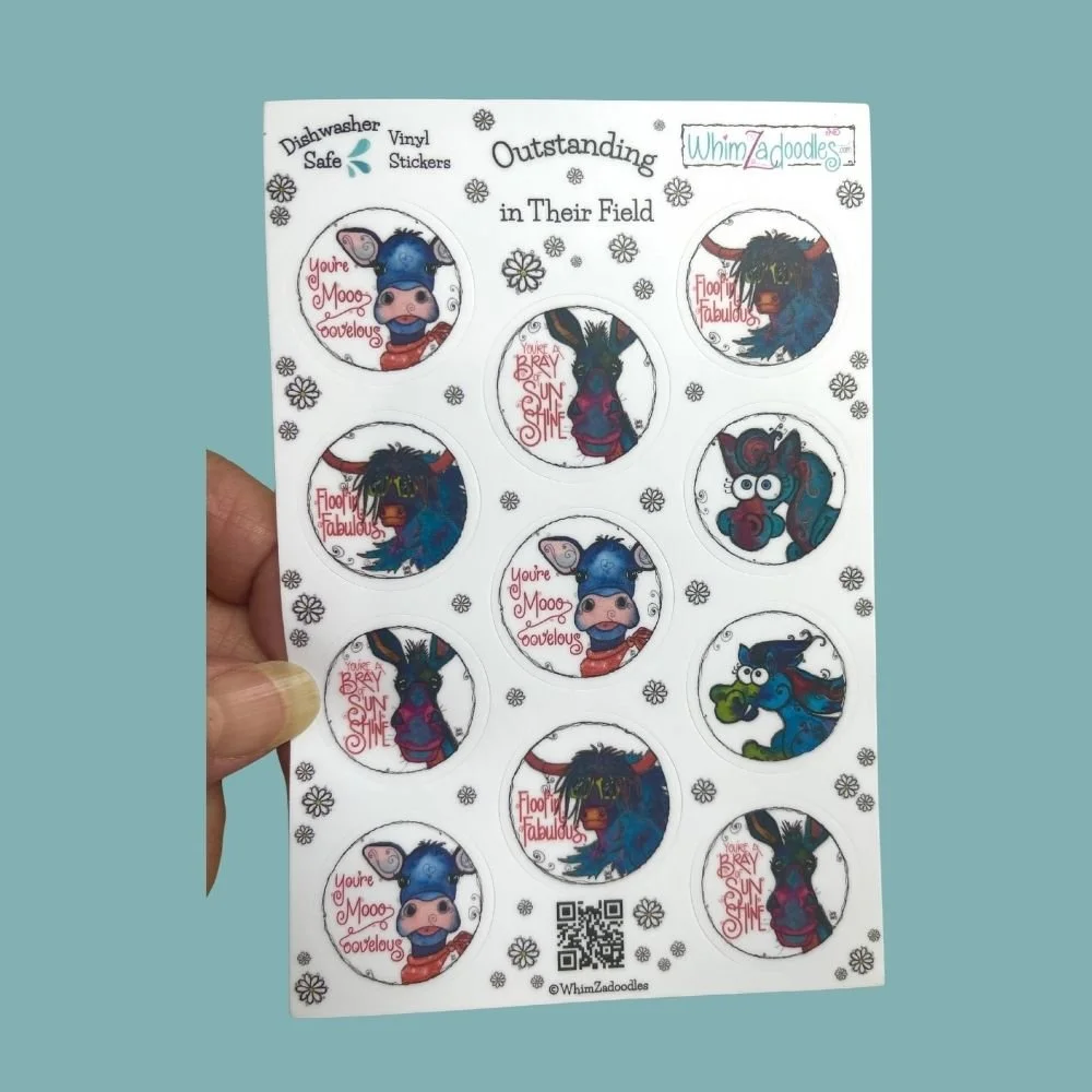 Hand holding the Outstanding in Their Field sticker sheet showing 11 colorful round WhimZaCritter stickers for scale.
