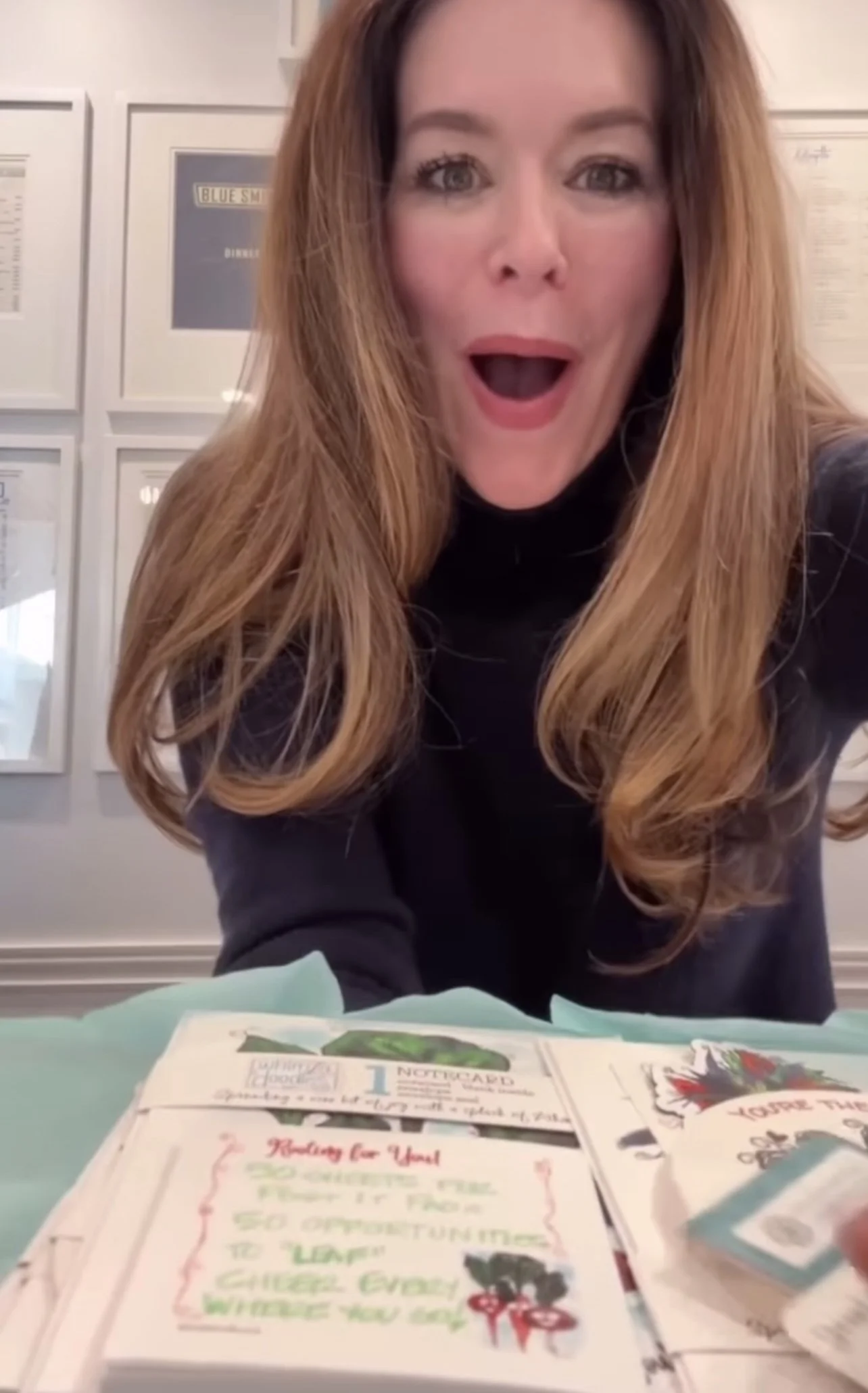 Colleen Williams reacting with surprise and delight while opening a WhimZadoodles package, surrounded by colorful cards and small gifts on teal tissue paper.