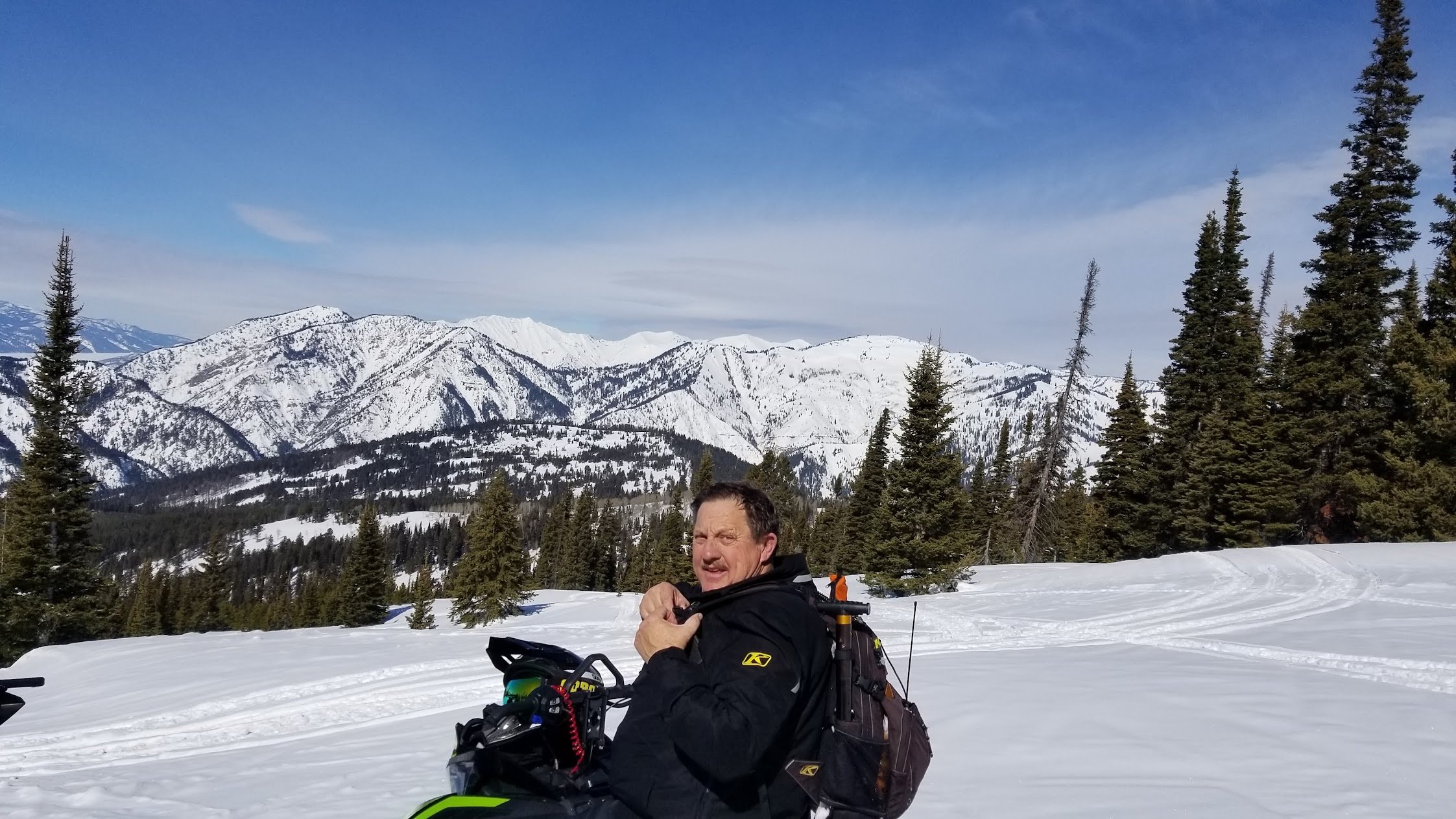 A man with a backpack sitting on a snowbike in a snowy mountain landscape with pine trees and snow-covered mountains in the background under a partly cloudy sky.