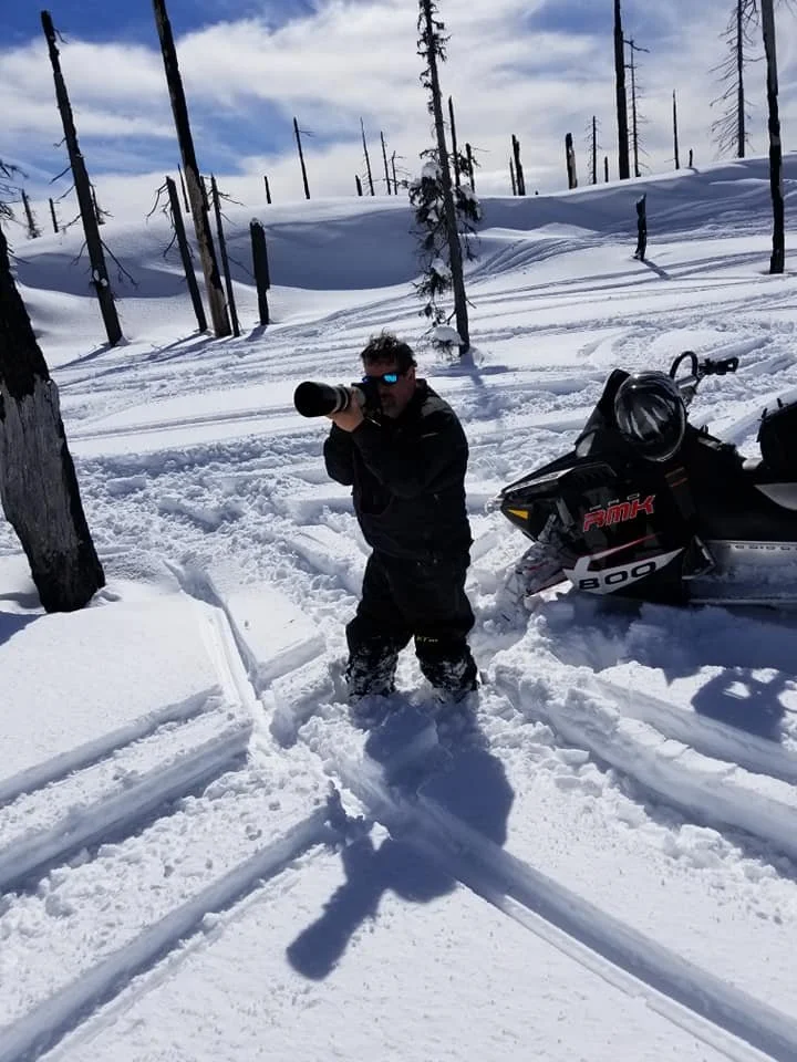 A person standing in snowy terrain, holding a camera, with a snowmobile nearby and a forest of leafless trees in the background under a partly cloudy sky.