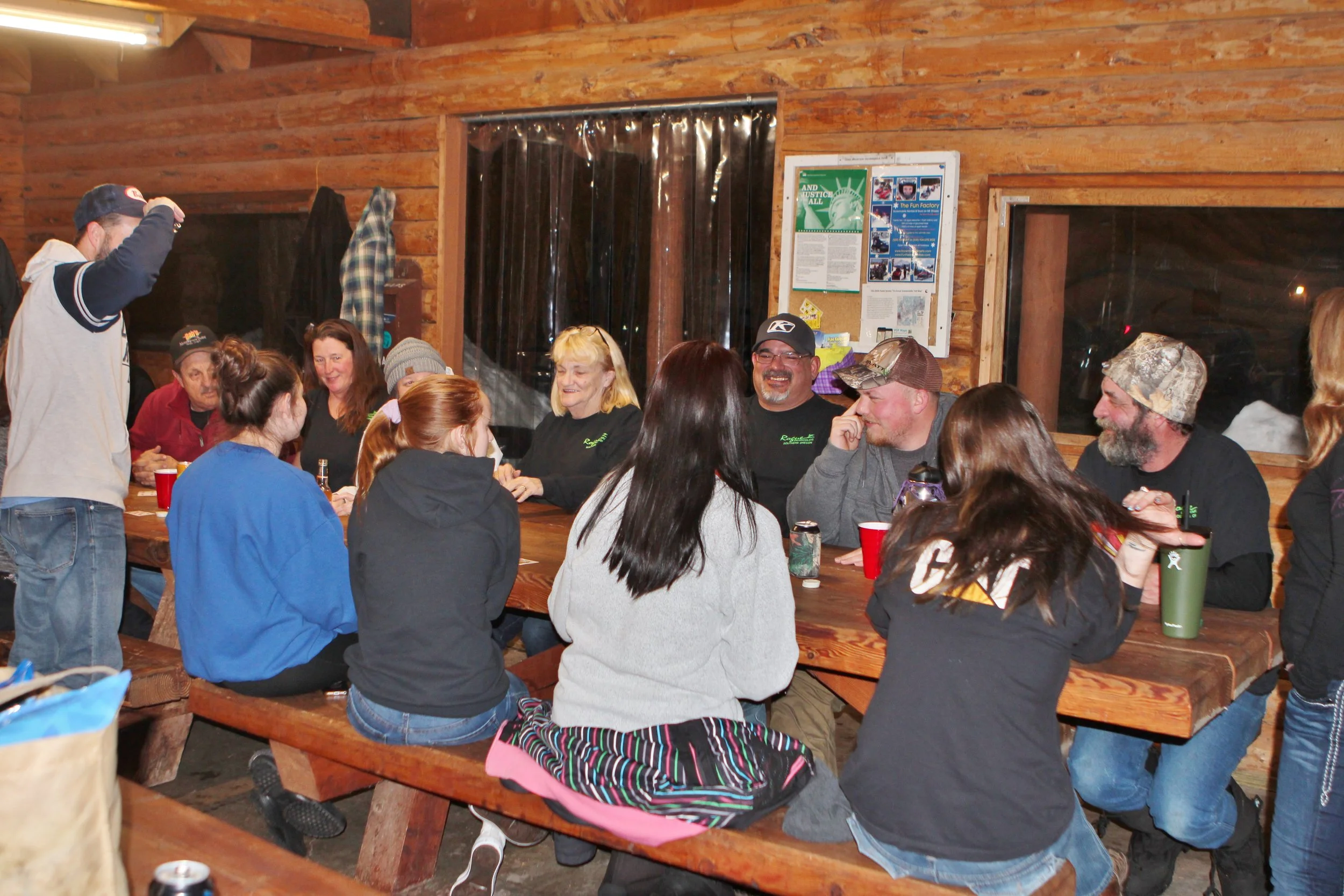A group of people sitting and standing around a long wooden table inside a rustic wooden cabin, enjoying a social gathering at night.