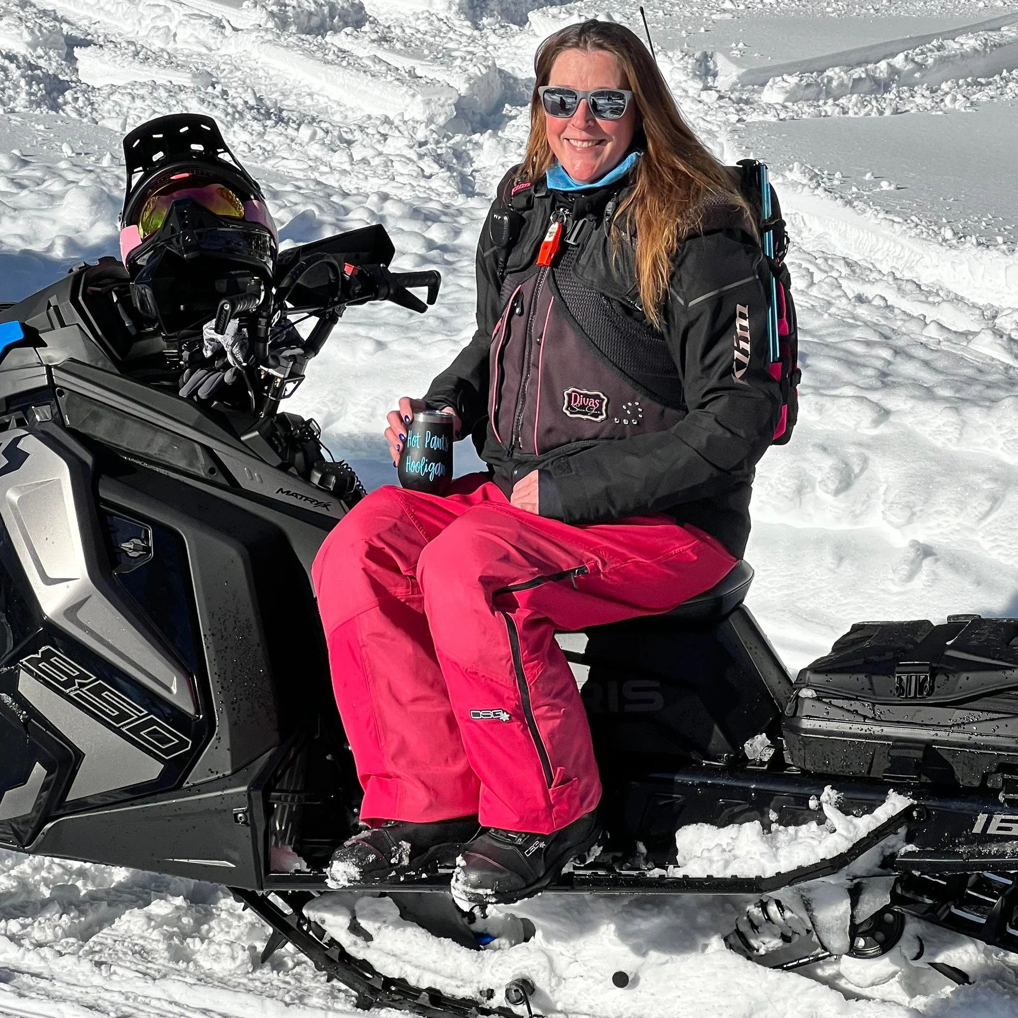 Woman in snow gear sitting on a snowmobile in snowy landscape, holding a mug that says "Hot Power Hooligan," smiling, with snow-covered ground around her.