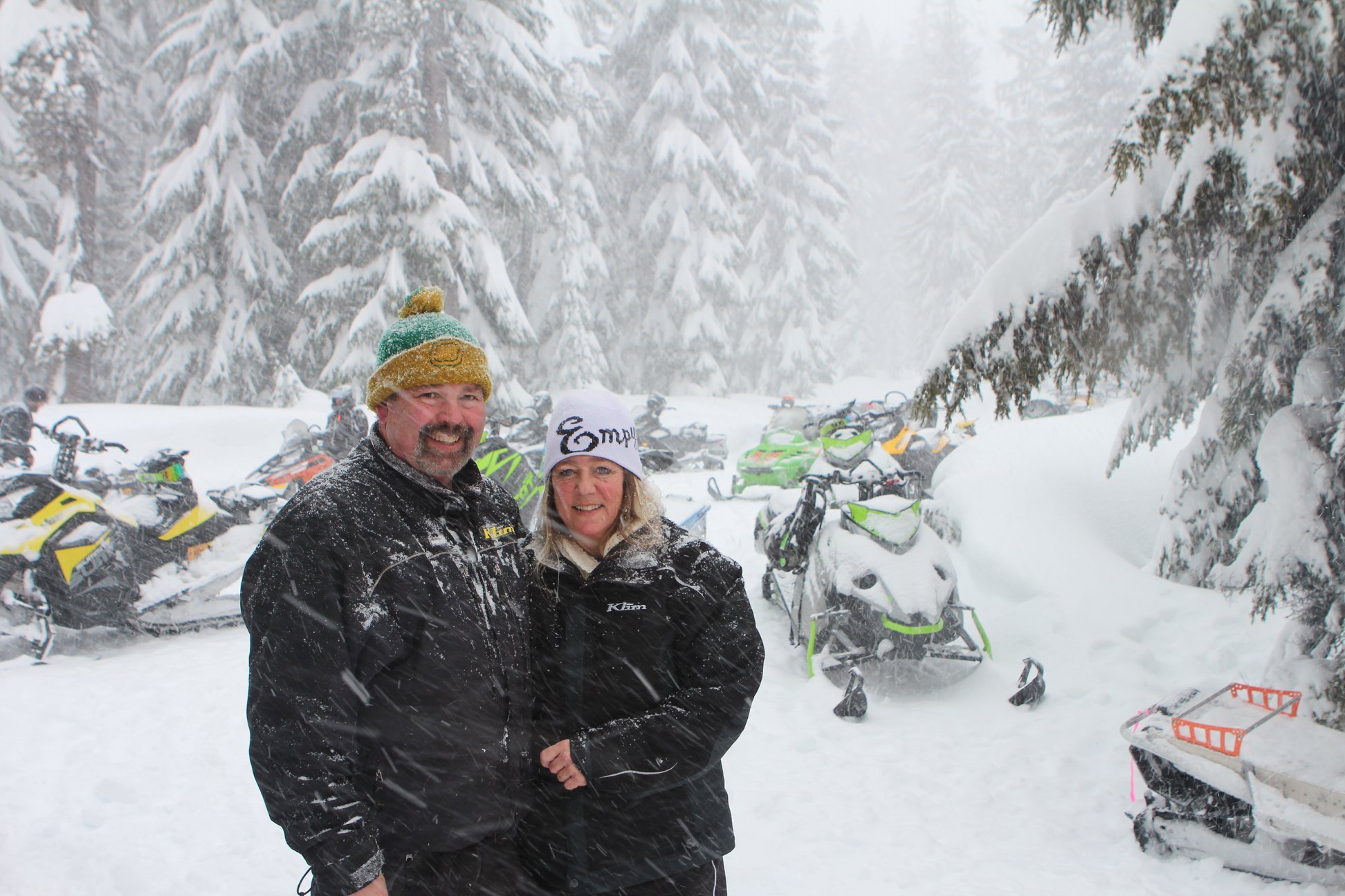 Two smiling people stand in a snowy forest with snowmobiles in the background.