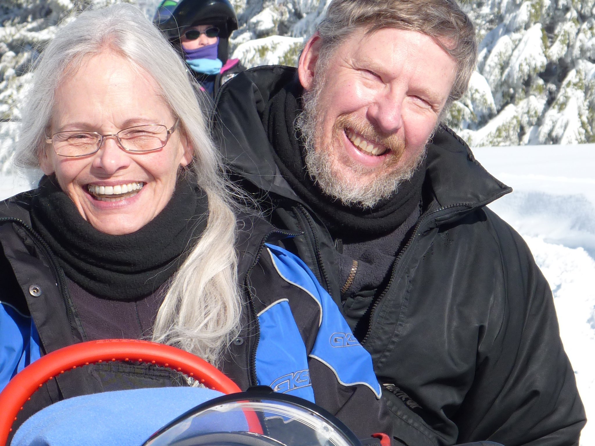 Smiling elderly woman with glasses and long gray hair, and smiling middle-aged man with a beard, both dressed warmly for snowy weather, with snow-covered trees in the background.