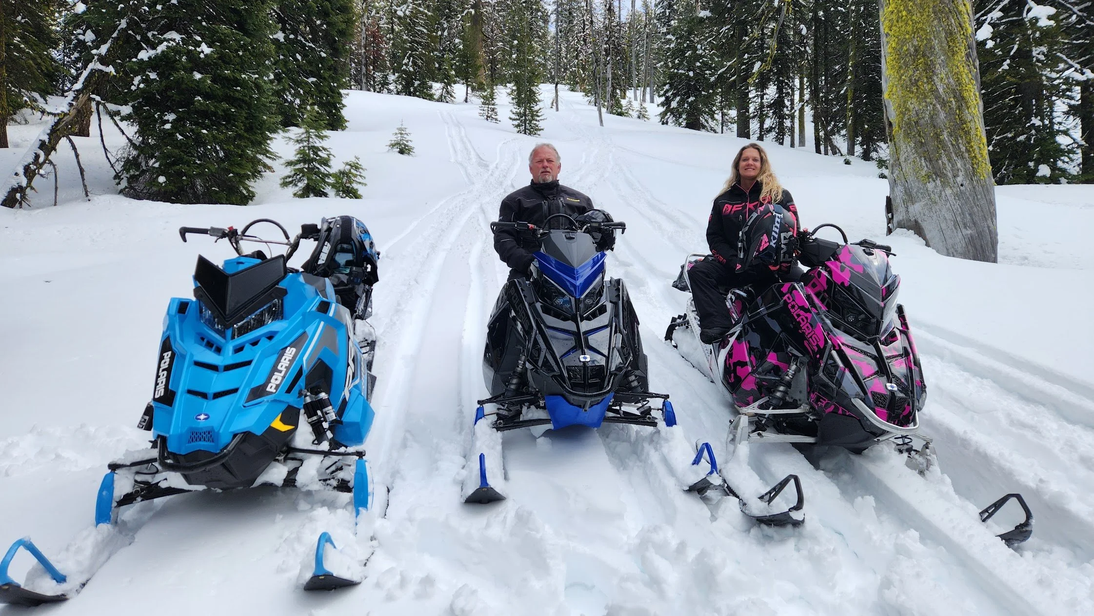 Two people, a man and a woman, sitting on colorful snowmobiles in a snow-covered forest clearing with tall pine trees.