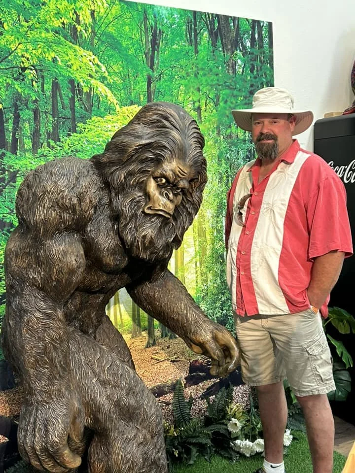 A man in a safari hat and casual pink and white shirt stands next to a large, realistic statue of a gorilla in front of a green forest background.