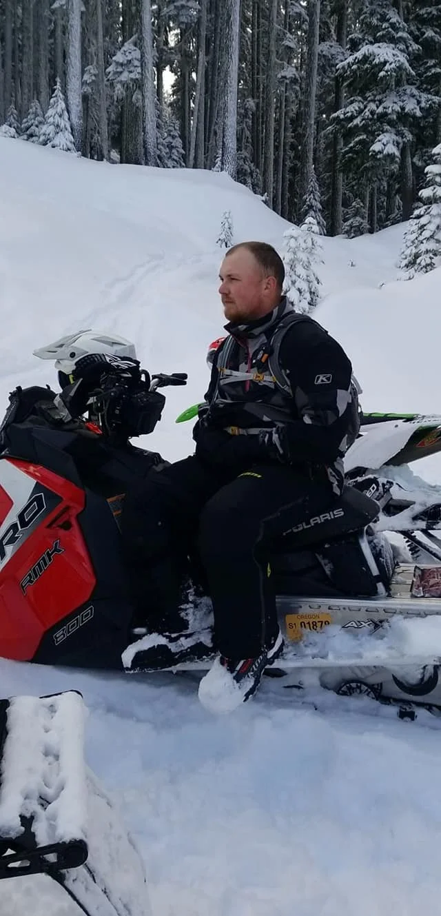 A man sitting on a red and black snowmobile in a snowy forest, wearing winter gear.