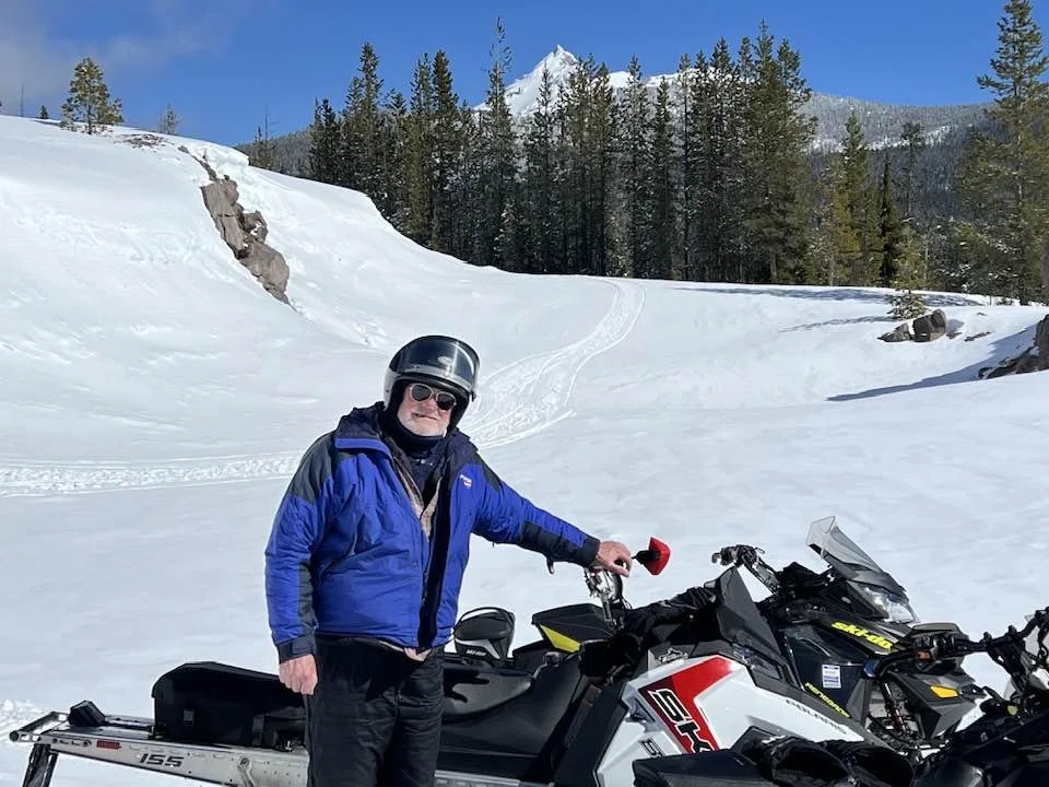 Man wearing sunglasses and a blue winter jacket standing next to snowmobiles on snowy terrain with pine trees and mountains in the background.