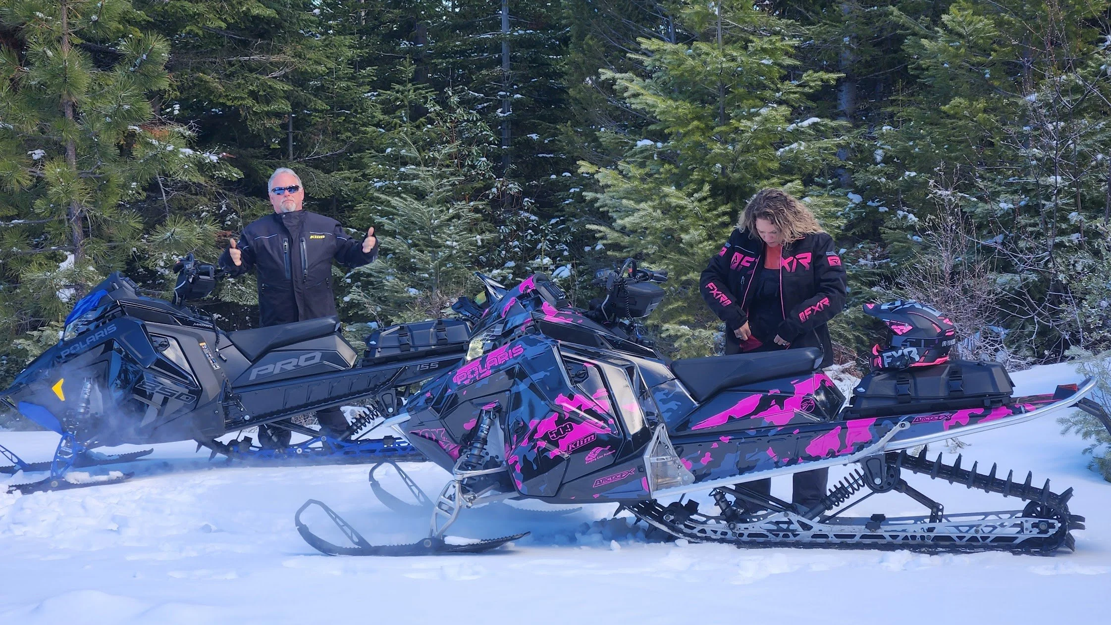 Two people with a black and pink snowmobile in a snowy forest, one man standing with thumbs up, wearing sunglasses and a black jacket, the woman looking down adjusting her jacket, both dressed in black and pink gear.