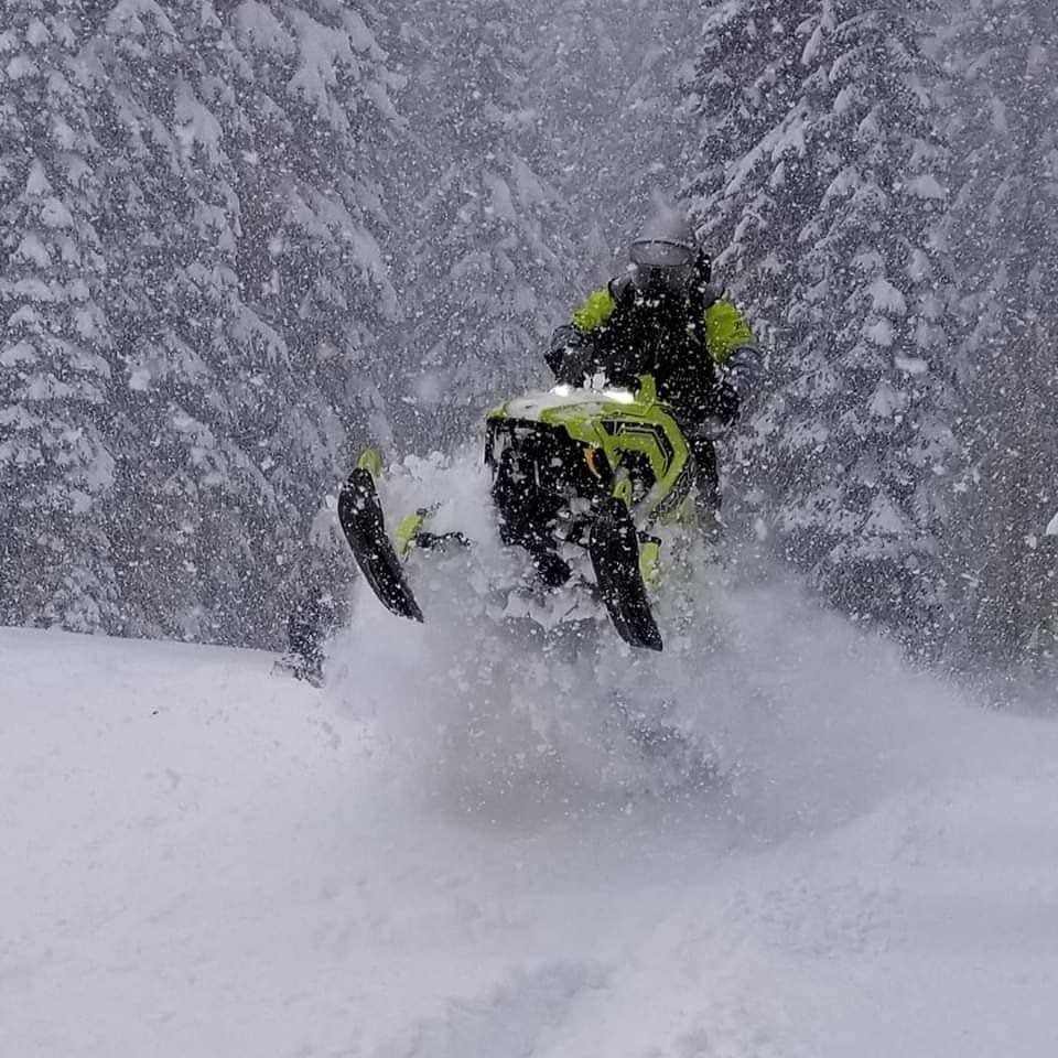 A person in bright yellow winter gear and a helmet riding a snowmobile through deep snow in a snowy forest.