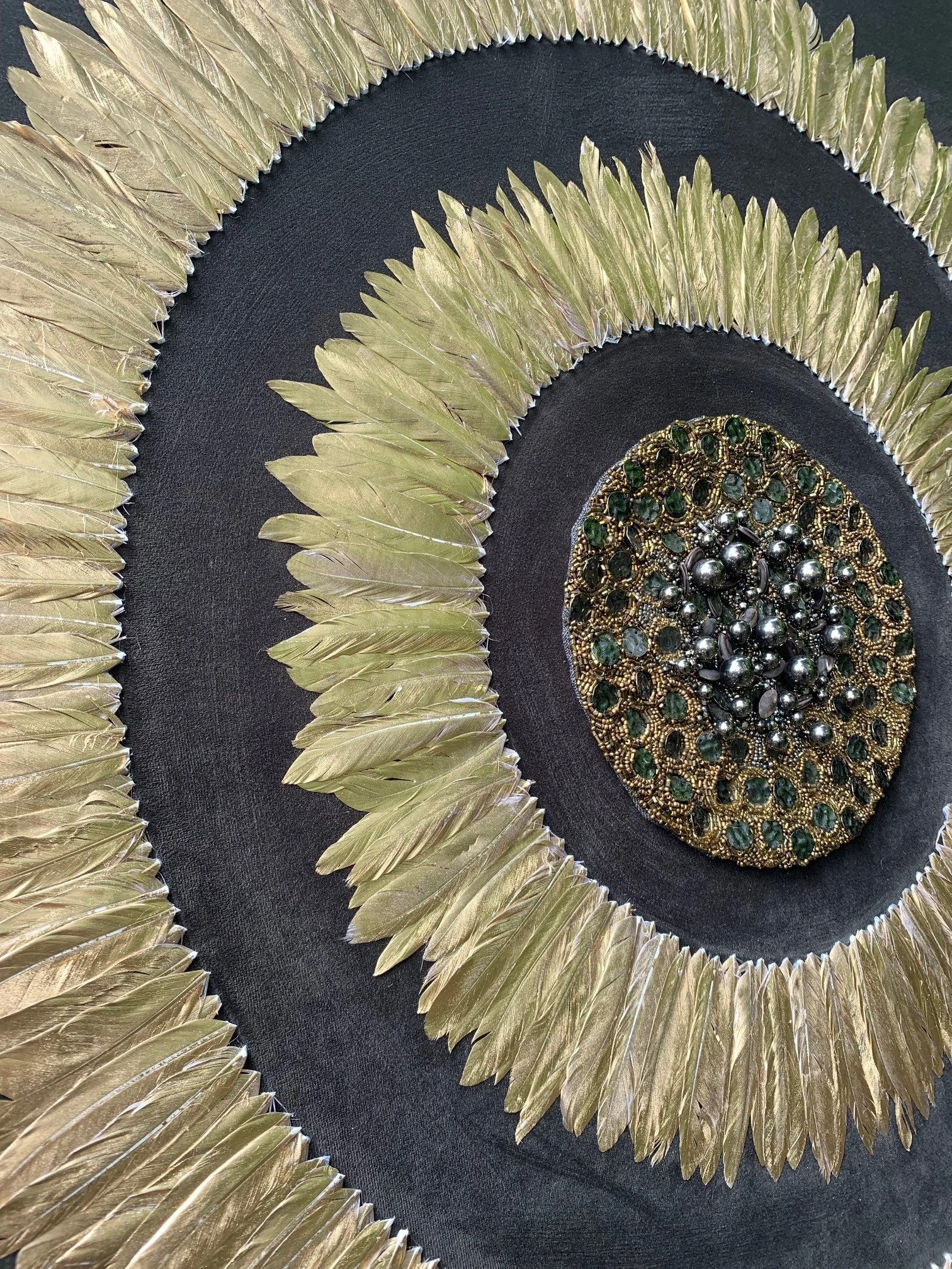 A decorative circular art piece featuring a central beaded and gemstone design, surrounded by gold-colored metallic feathers on a black background.