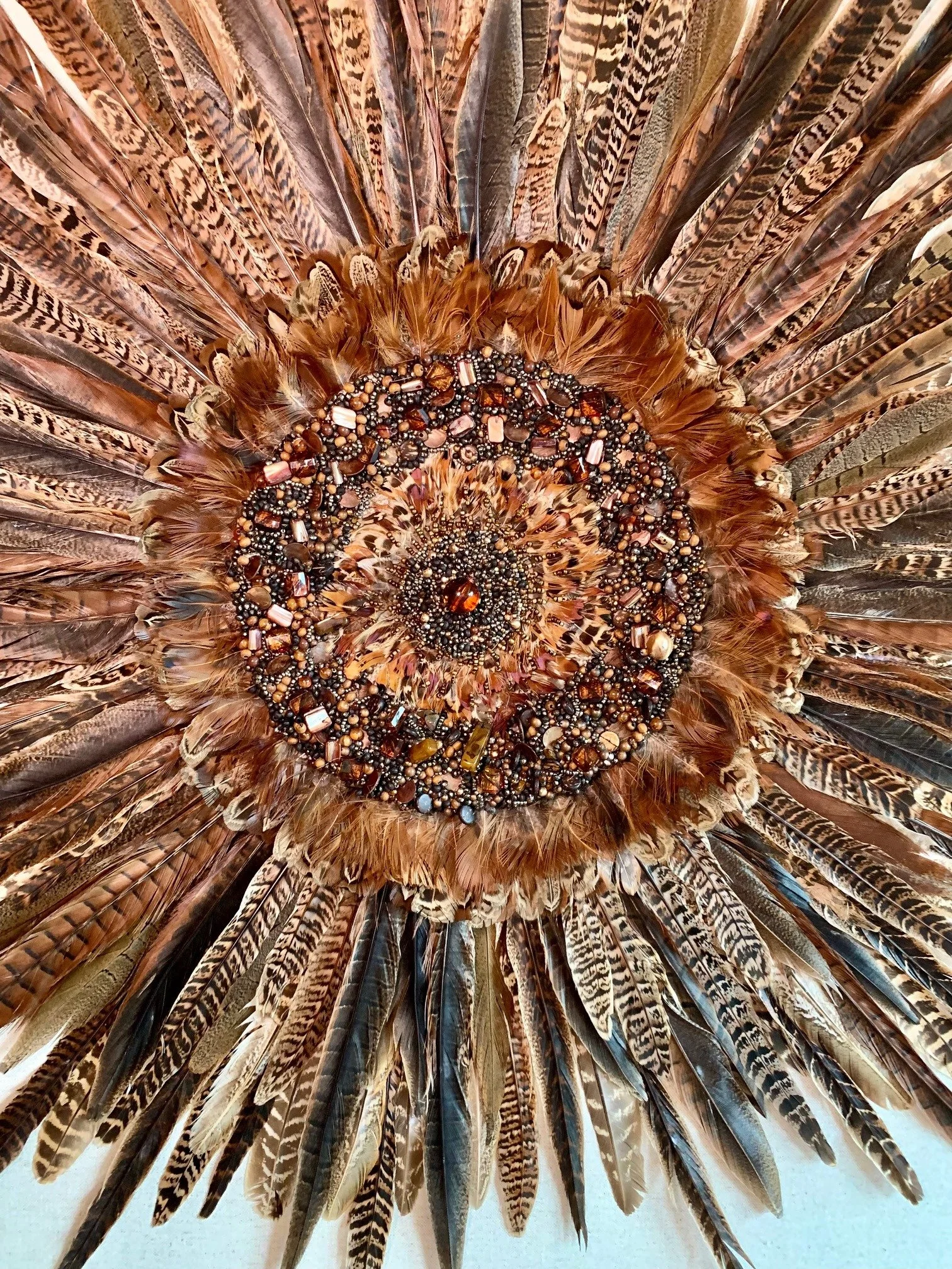 A decorative display made from a large collection of feathers arranged in a circular pattern, adorned with various small beads and stones in the center.
