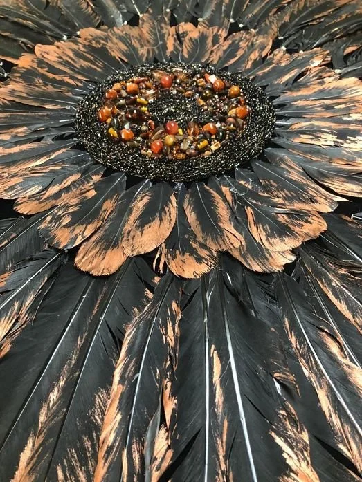 A decorative arrangement resembling a sunflower with black and tan feathers, featuring a center filled with small beads and round objects.