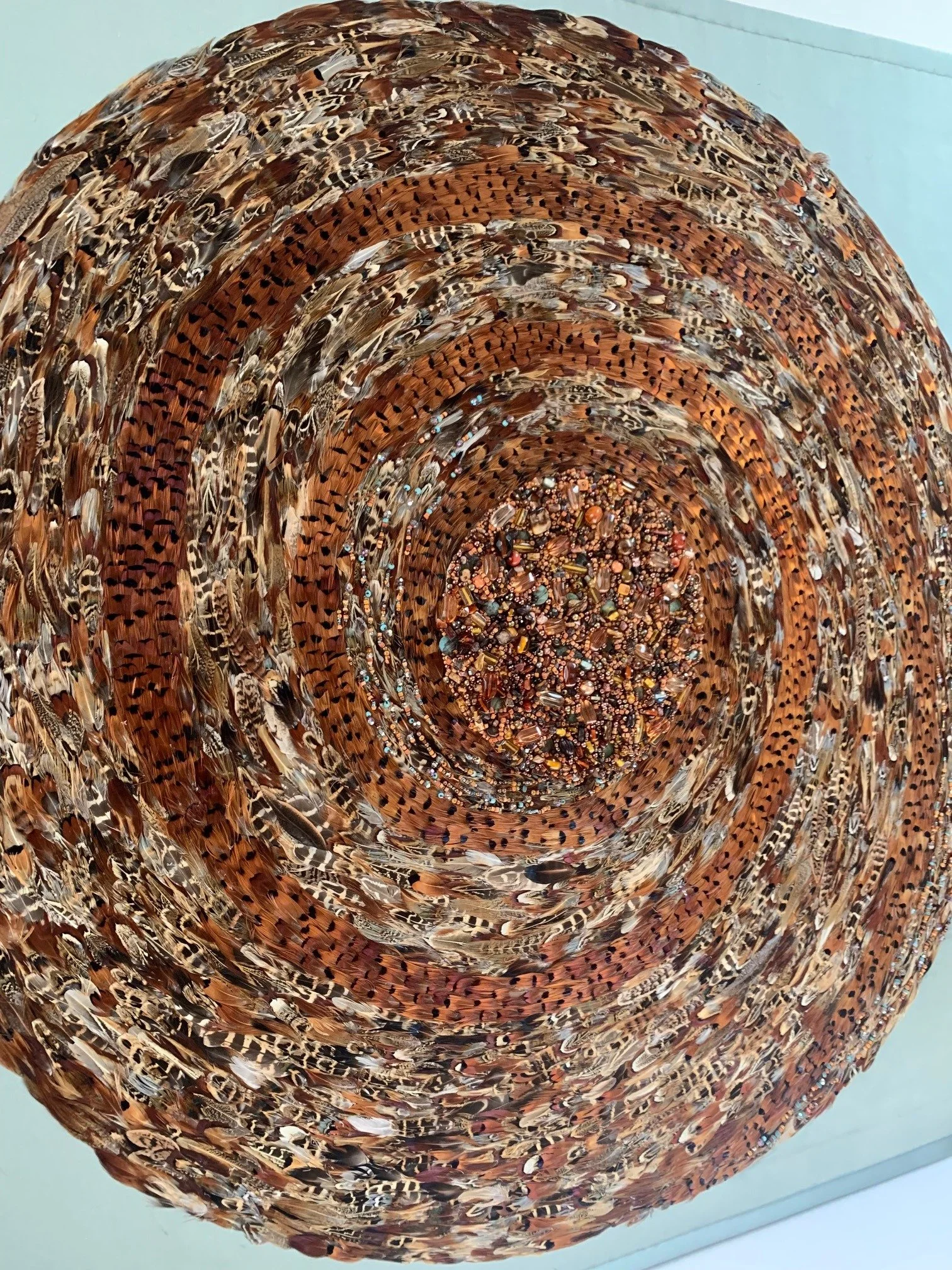 A large, circular piece of artwork made from layered bird feathers arranged in a spiral pattern.