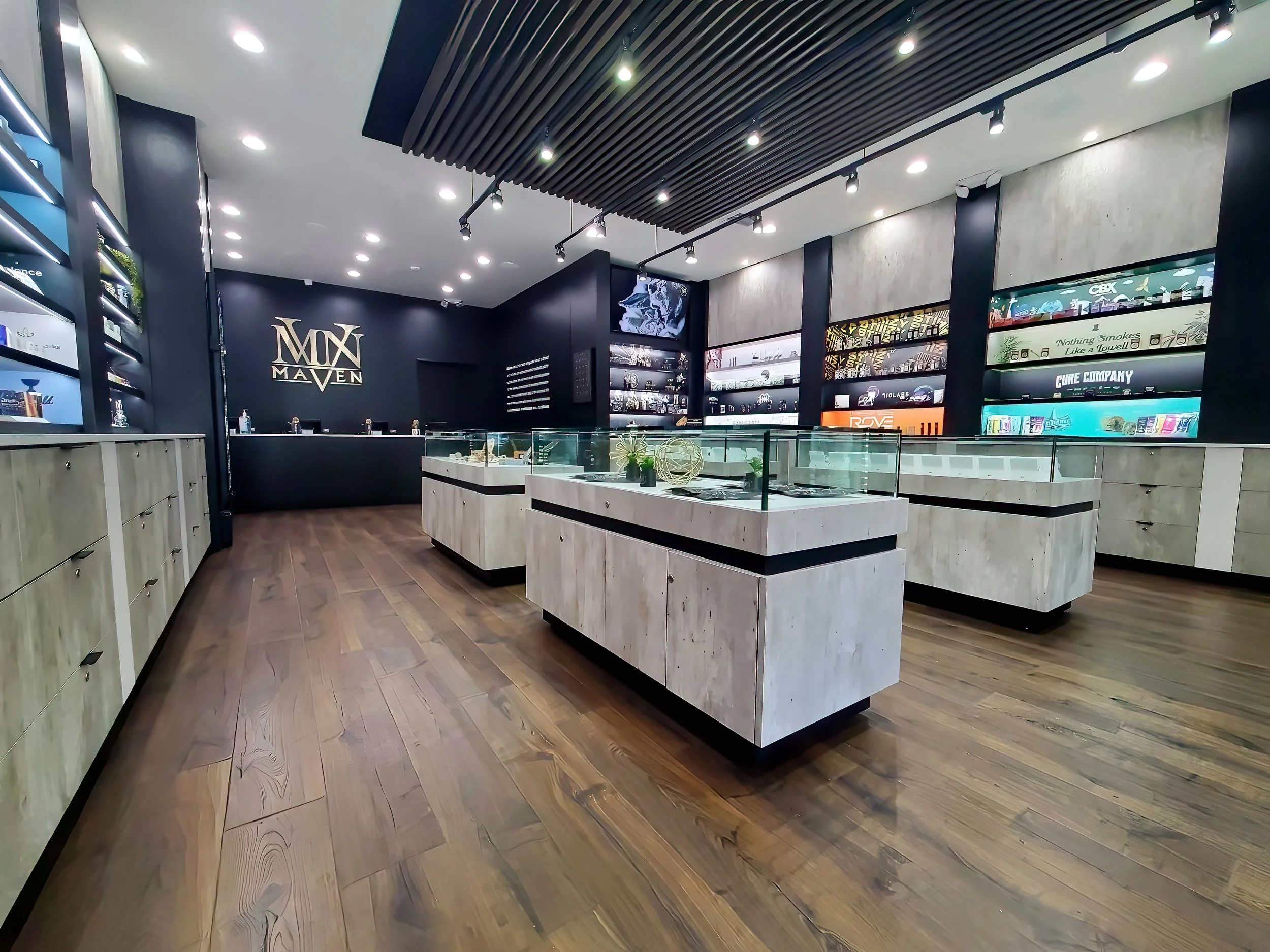 Interior of a modern store with black walls, wooden flooring, glass display cases, and shelves with various products, featuring the Maven logo on the wall in gold.