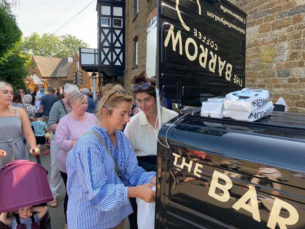 People lined up at a black food truck called 'The Barrow Coffee' in a neighborhood setting, with a brick building and trees in the background.