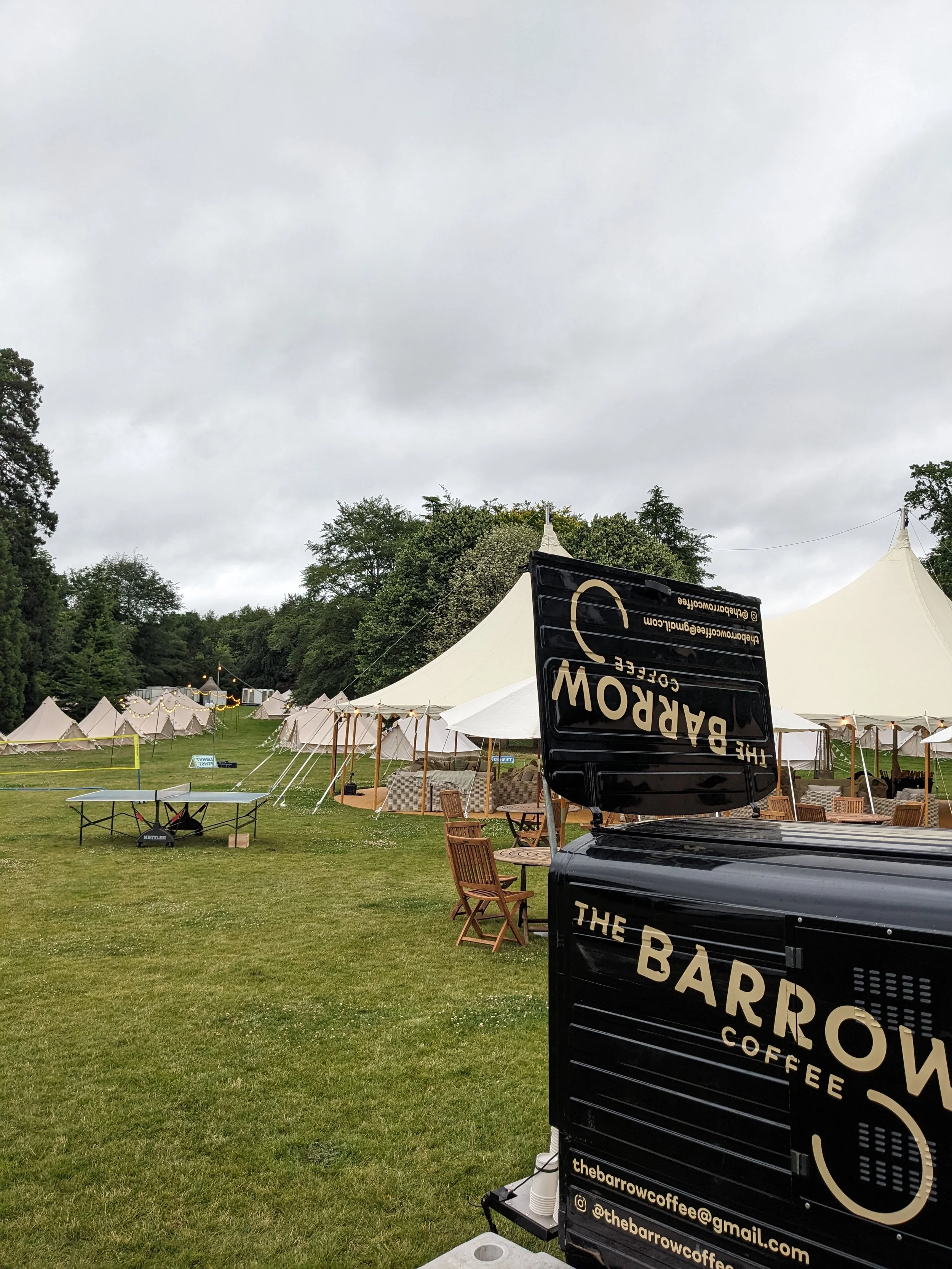 Outdoor event setup with white tents, wooden chairs, and a black coffee trailer labeled 'The Barrow Coffee' on a grassy field under a cloudy sky.