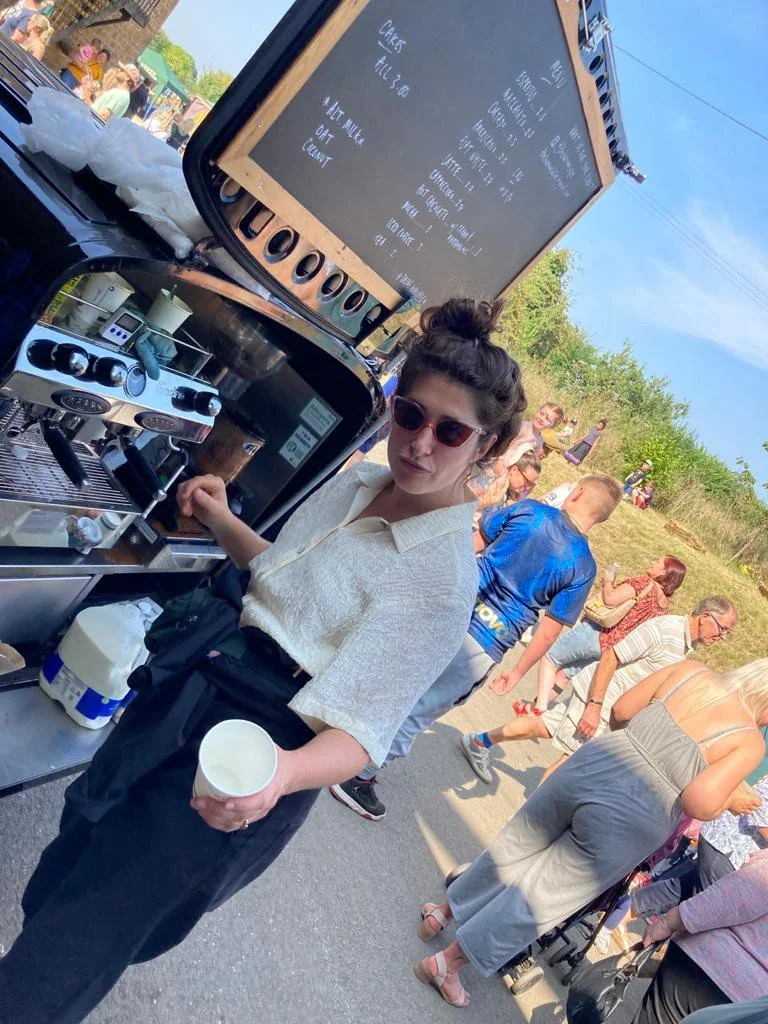 A young woman with dark hair in a bun wearing sunglasses, a beige short-sleeve shirt, and black pants, making coffee using a professional espresso machine at an outdoor market. She holds a white cup and stands near a black coffee cart. People are wal