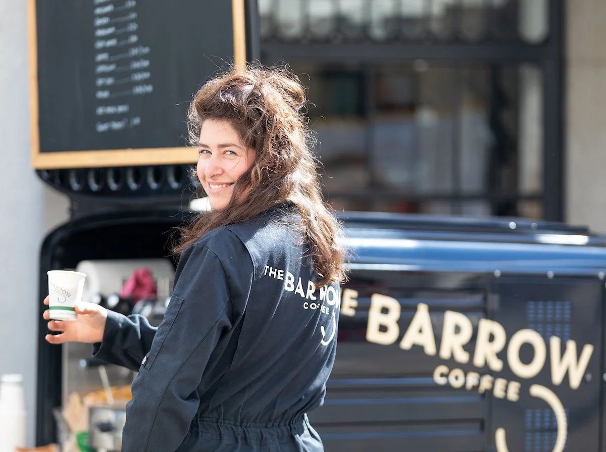 A woman with curly brown hair in a dark jacket with a logo that reads 'The Barrow Coffee' on the back, smiling and holding a cup of coffee in front of a black coffee truck with the samelogo.