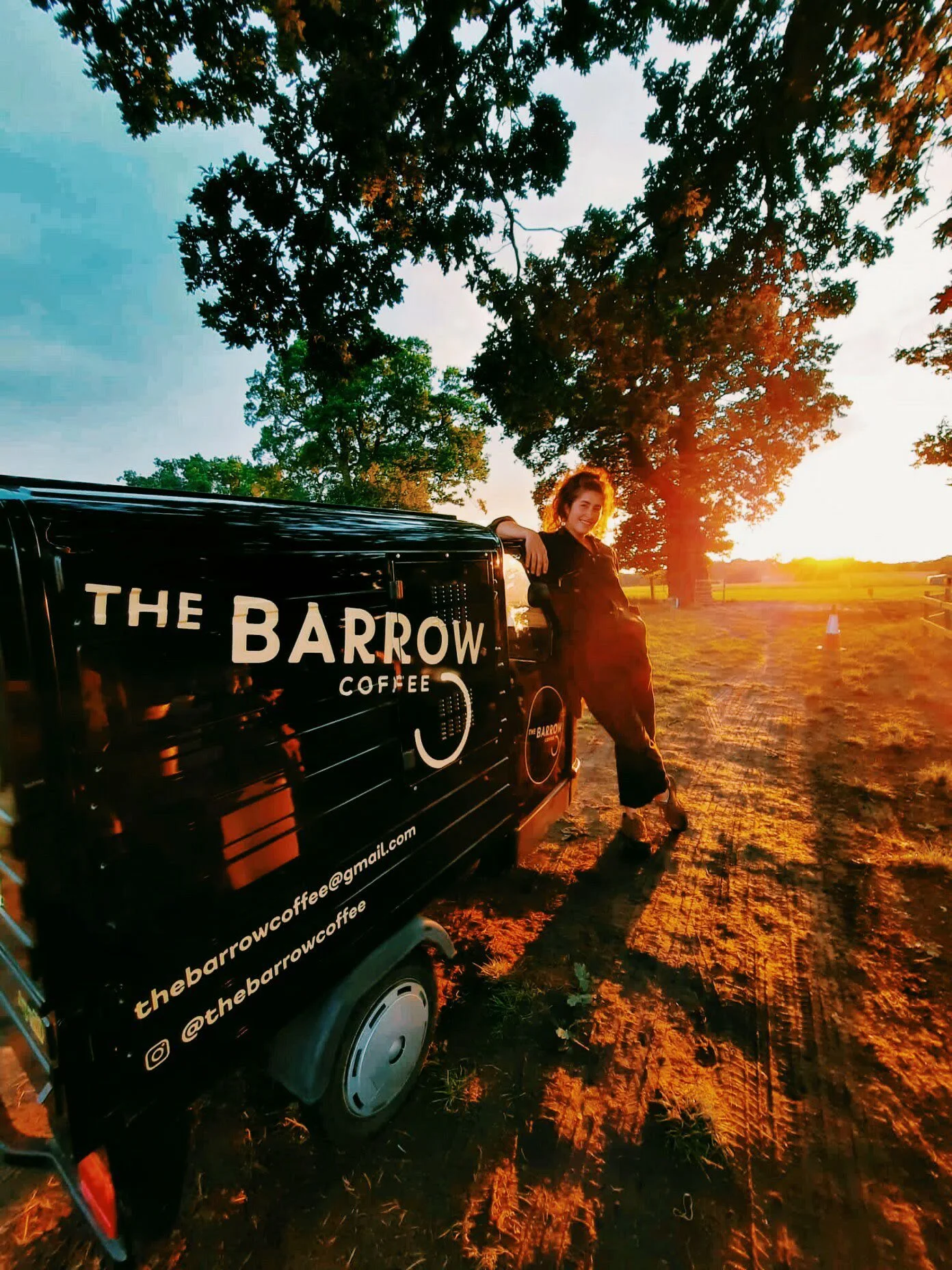 A woman leaning on a black coffee cart with 'The Barrow Coffee' logo, standing outdoors during sunset with tall trees and a dirt path in the background.