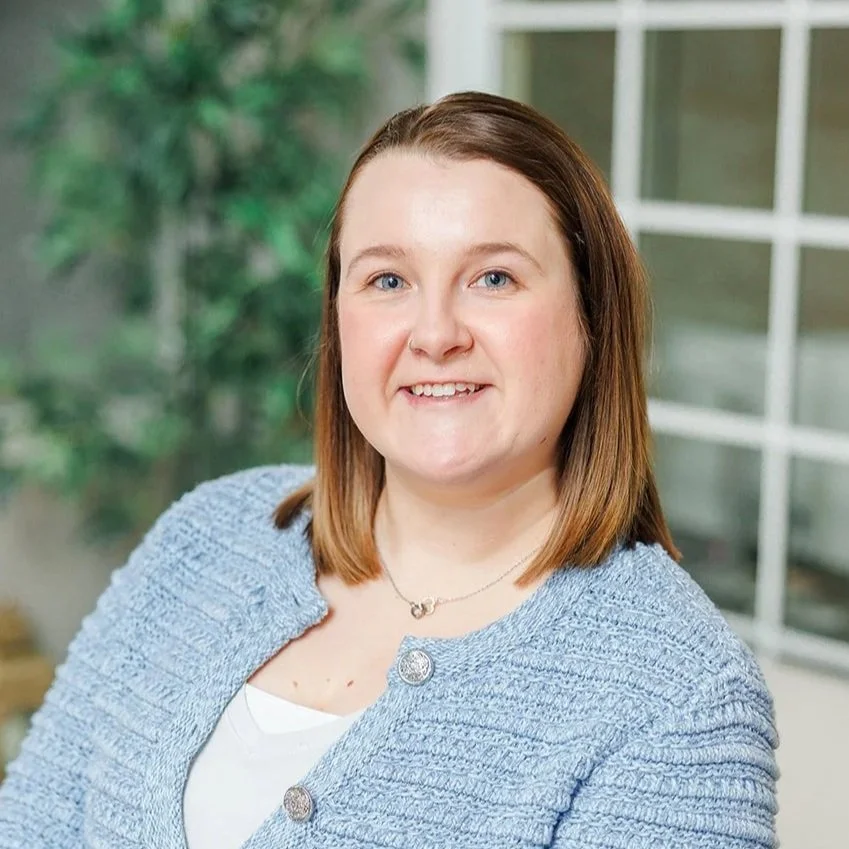 A woman with long brown hair, wearing a bright orange blouse with a zippered neckline, standing indoors with a blurred background featuring wooden and metal elements.