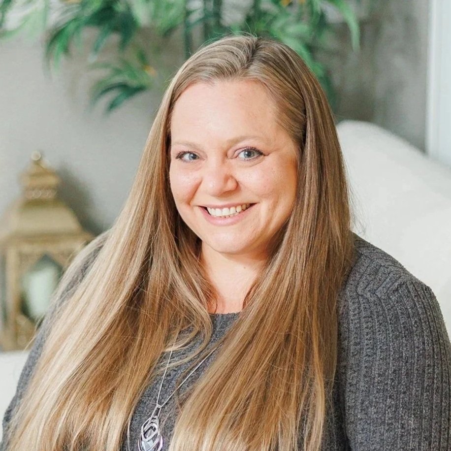 A woman with long, wavy brown hair, smiling, wearing a gray jacket and a navy blue top, with a necklace, standing in an indoor setting with artwork and furniture in the background.