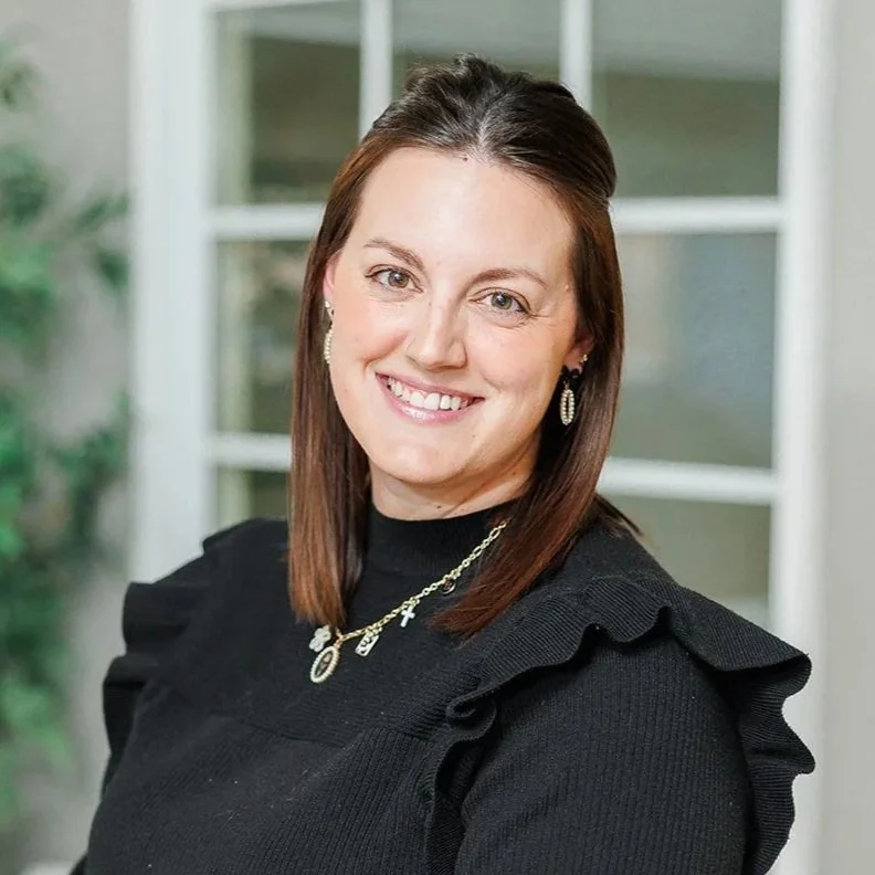A woman with shoulder-length dark brown hair, wearing a tan blazer over a red patterned blouse, smiling at the camera.