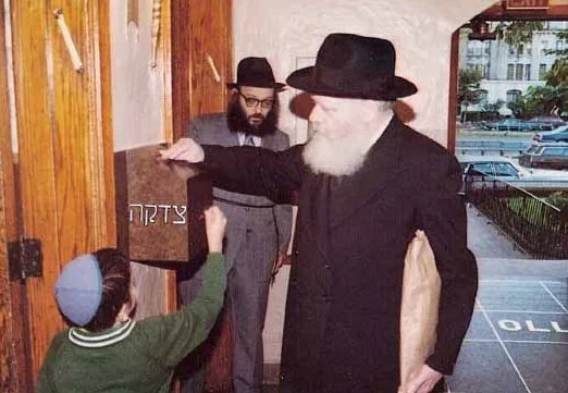 The Rebbe places a coin in a tzedakah box